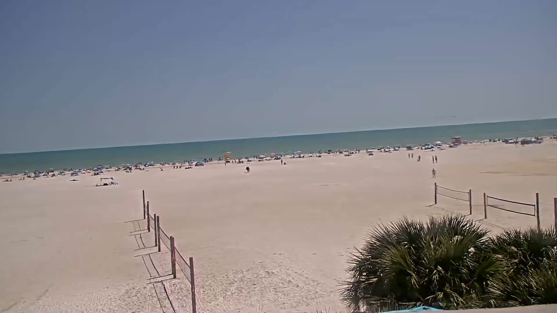 A wide sandy beach with scattered people and volleyball nets is visible, under a clear blue sky, with a calm, teal ocean in the background.