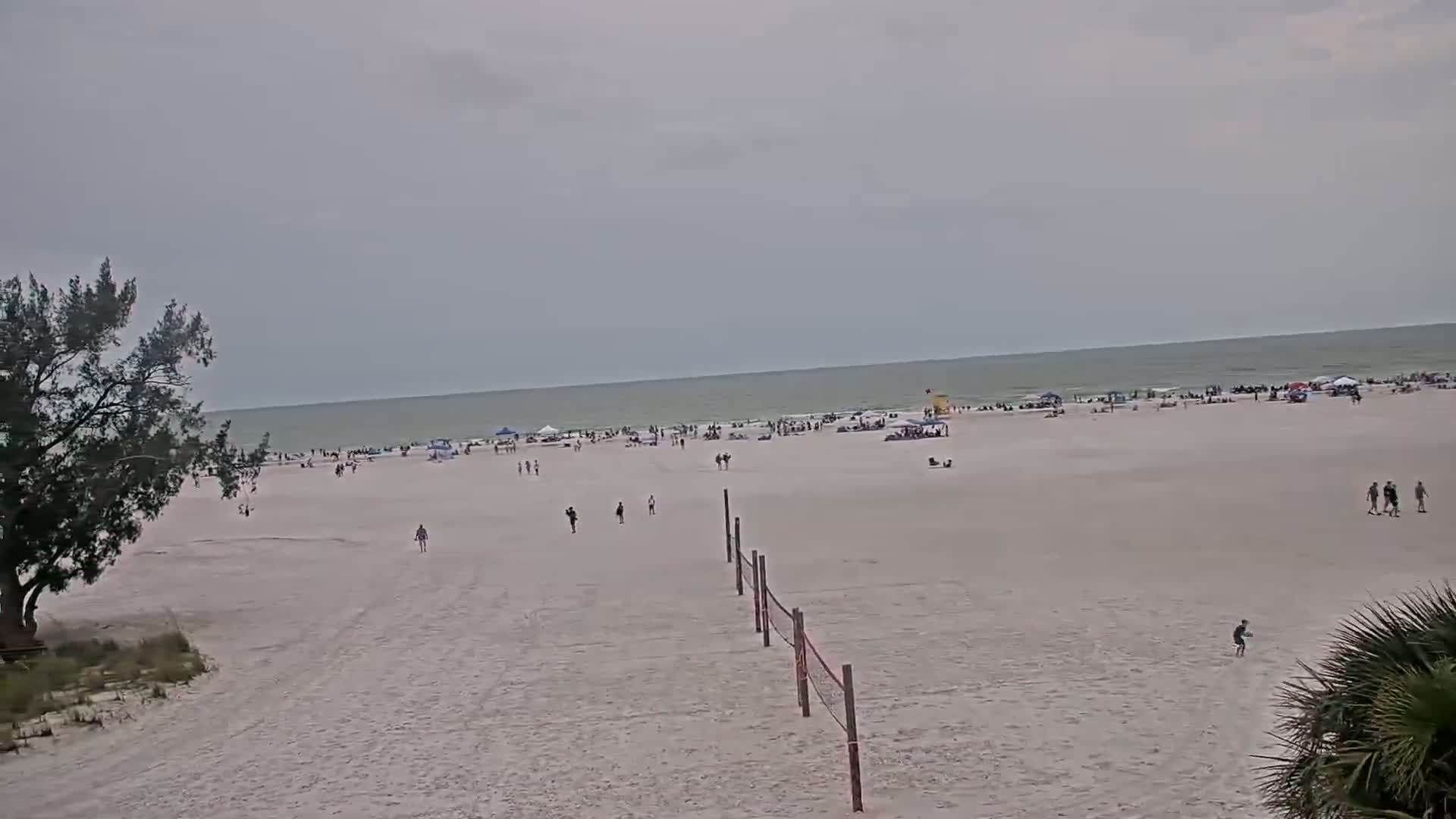 A wide sandy beach with scattered people, a volleyball net, and a calm, overcast ocean stretches to the horizon.