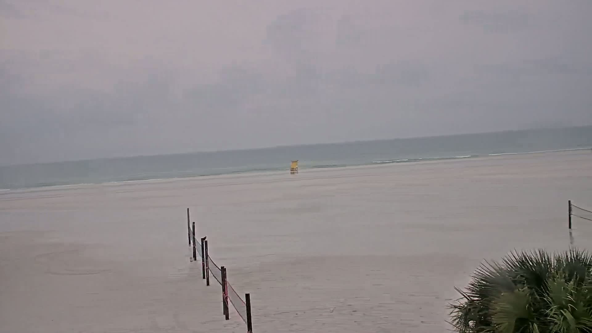 A mostly overcast beach scene shows a calm ocean, flooded sand, and volleyball nets partially submerged in the water.
