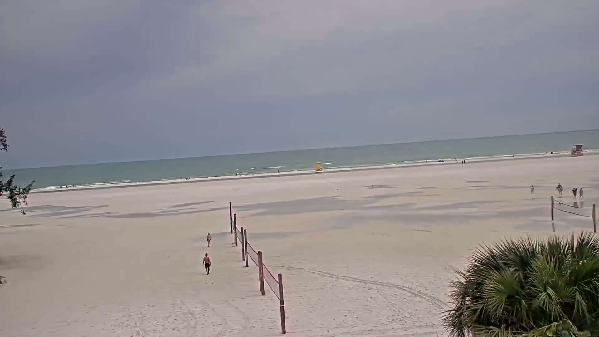 A wide expanse of sandy beach stretches to a calm, light-green ocean under a mostly cloudy sky, with a few people scattered across the beach and volleyball nets in the foreground.