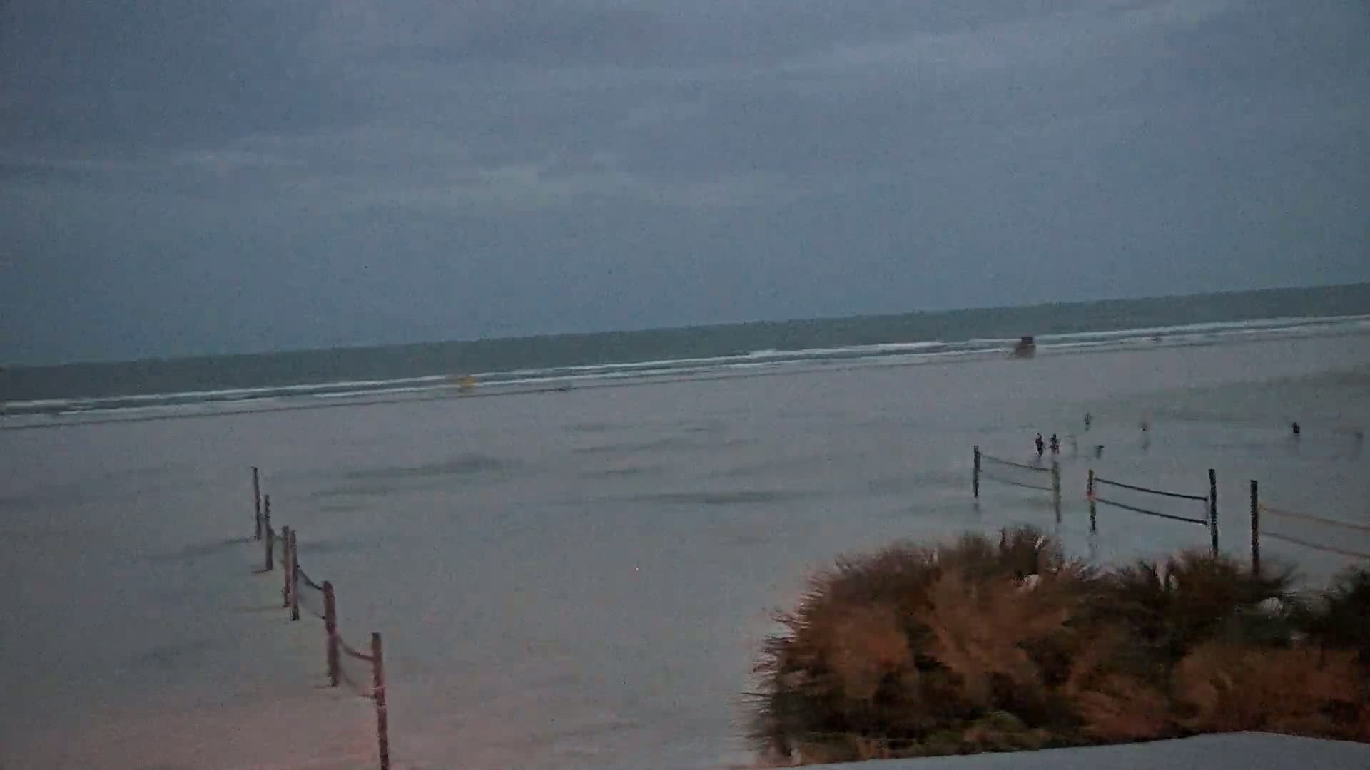 A wide, flat beach with volleyball nets and a few people is visible under an overcast sky, with a calm ocean in the background.