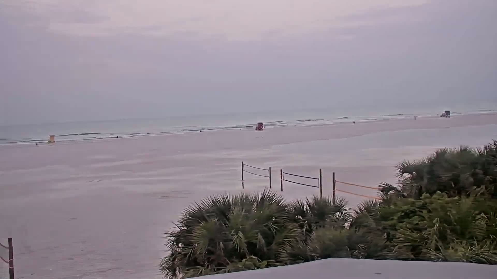 A mostly overcast beach scene shows a wide expanse of wet sand with several lifeguard stands and volleyball nets, partially submerged in shallow water,  behind a line of palm trees.