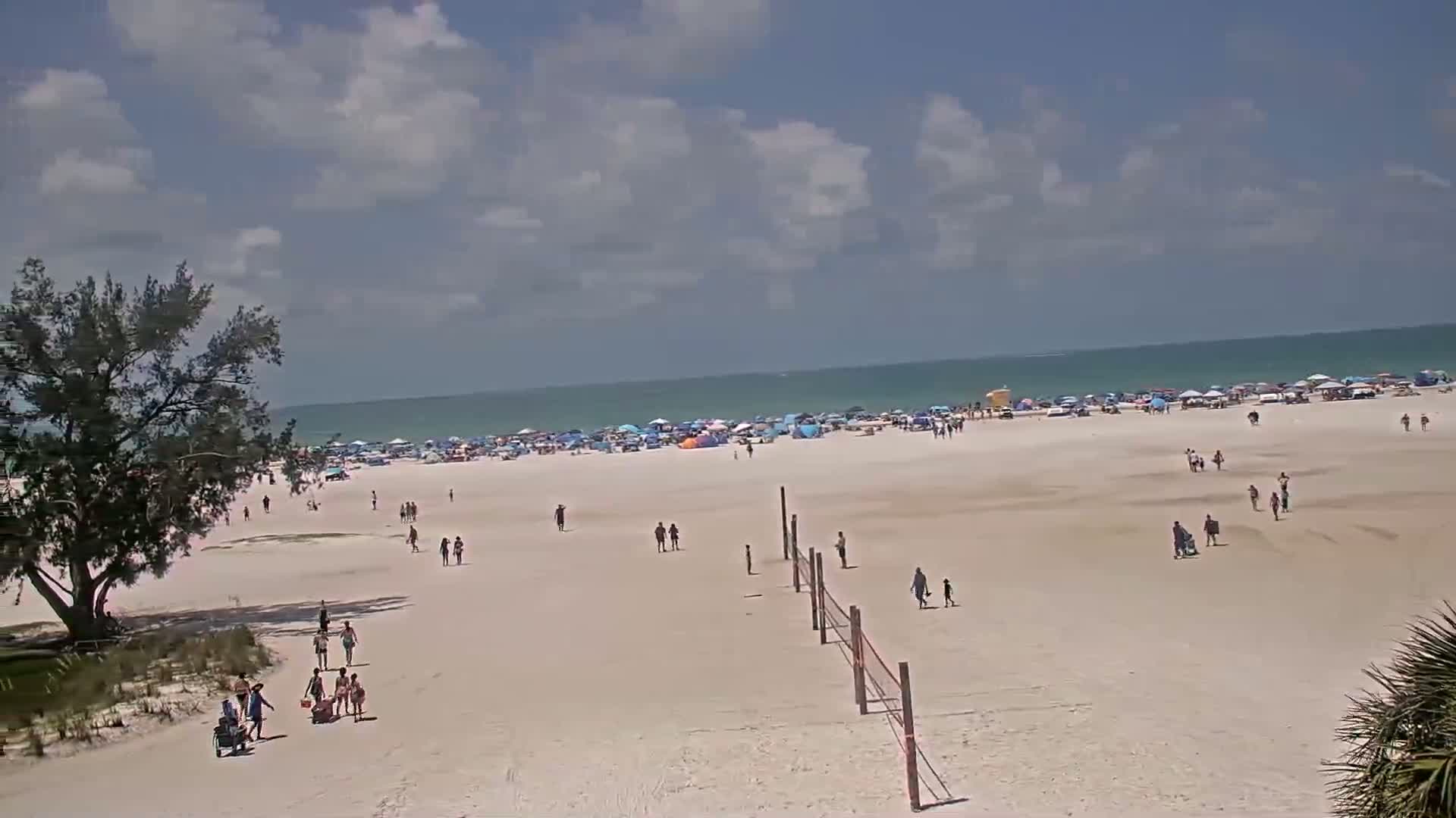 A mostly sunny day at a crowded beach shows people scattered across the sand with many umbrellas and beach equipment near the water's edge.