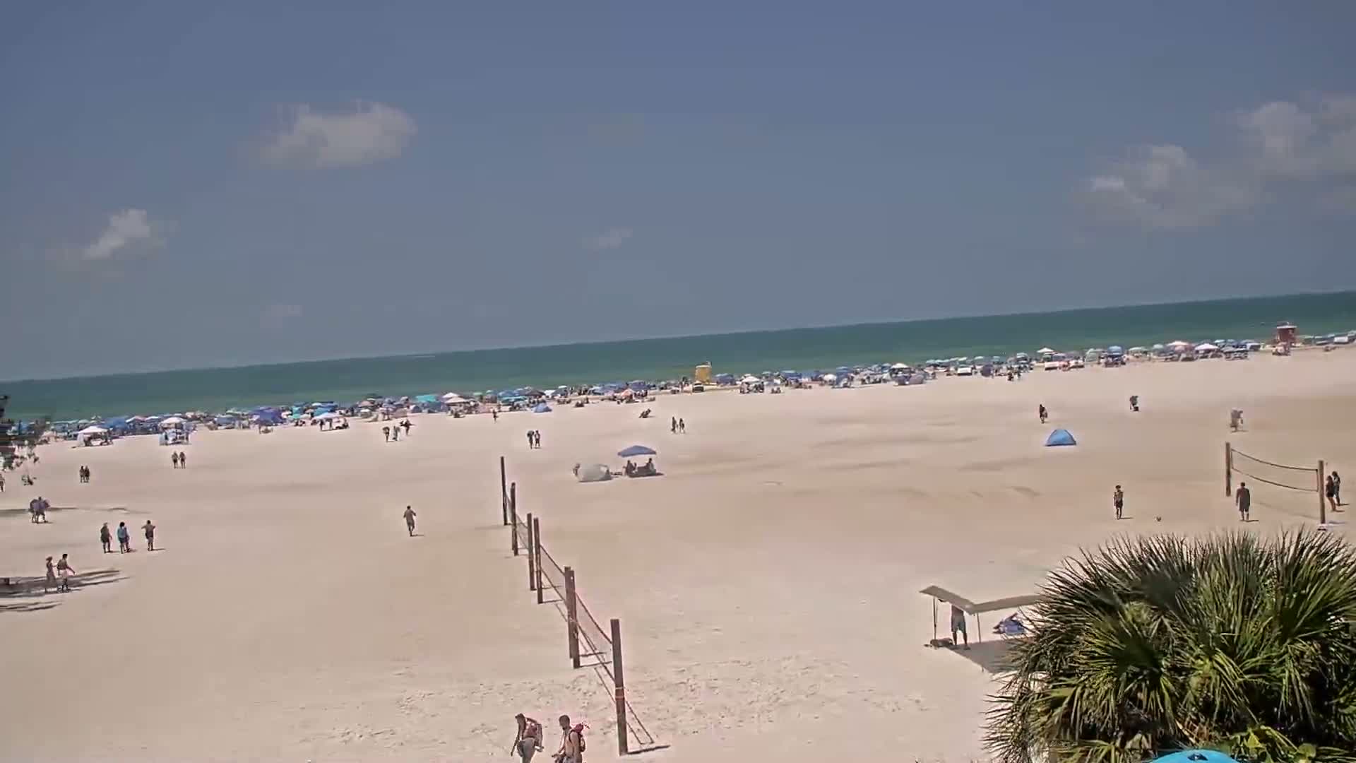 A sunny day at a crowded beach shows many people and umbrellas on the sand with a volleyball net and calm ocean in the background.