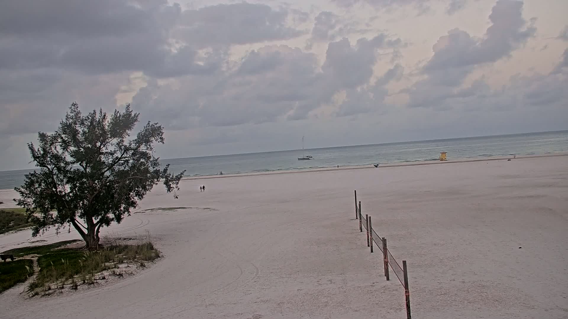 A mostly cloudy day on a white sand beach shows a lone tree near the dunes, a volleyball net, and a boat in the distance.