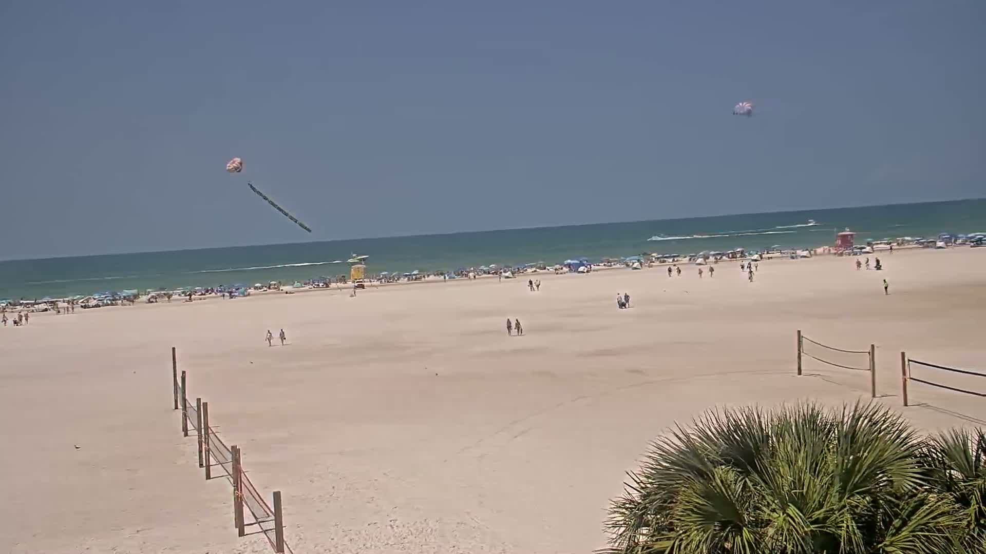 A sandy beach with scattered people, volleyball nets, and two parasailers in the sky under a clear blue sky.