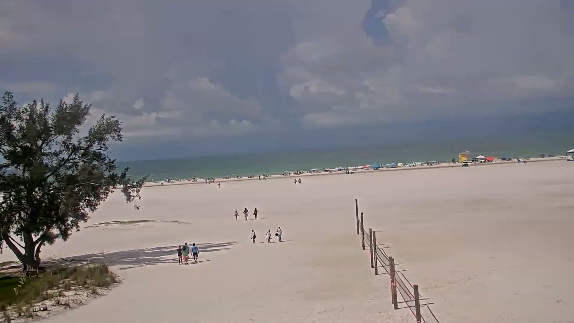 A mostly sunny beach scene shows a wide expanse of light-colored sand with scattered people, a few beach umbrellas near the water, and a partially cloudy sky.