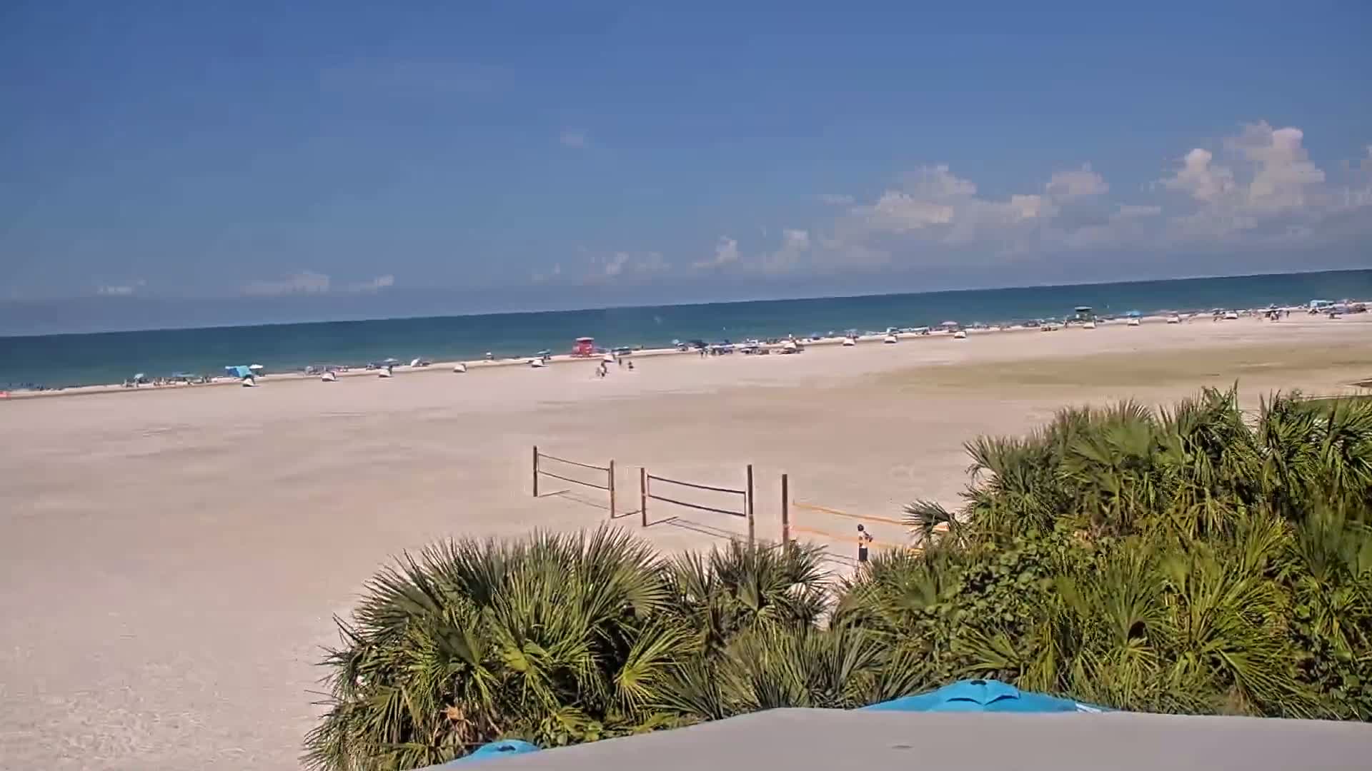A mostly sunny beach scene shows a wide expanse of light-colored sand with scattered people and beach umbrellas, volleyball nets, and some vegetation in the foreground, all under a mostly clear blue sky with a few clouds.