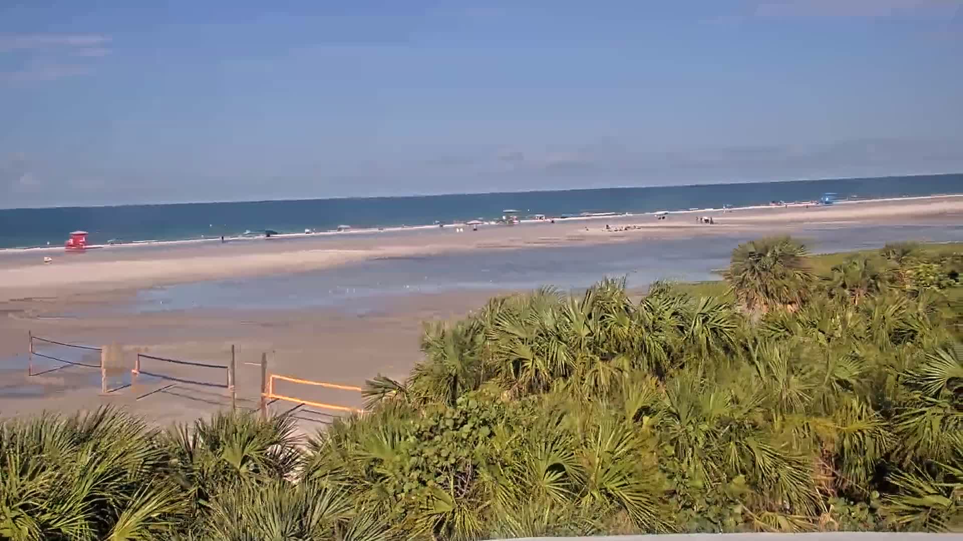 A beach scene on a sunny day shows a wide expanse of sand and shallow water with scattered people, volleyball nets, and lush green vegetation in the foreground.