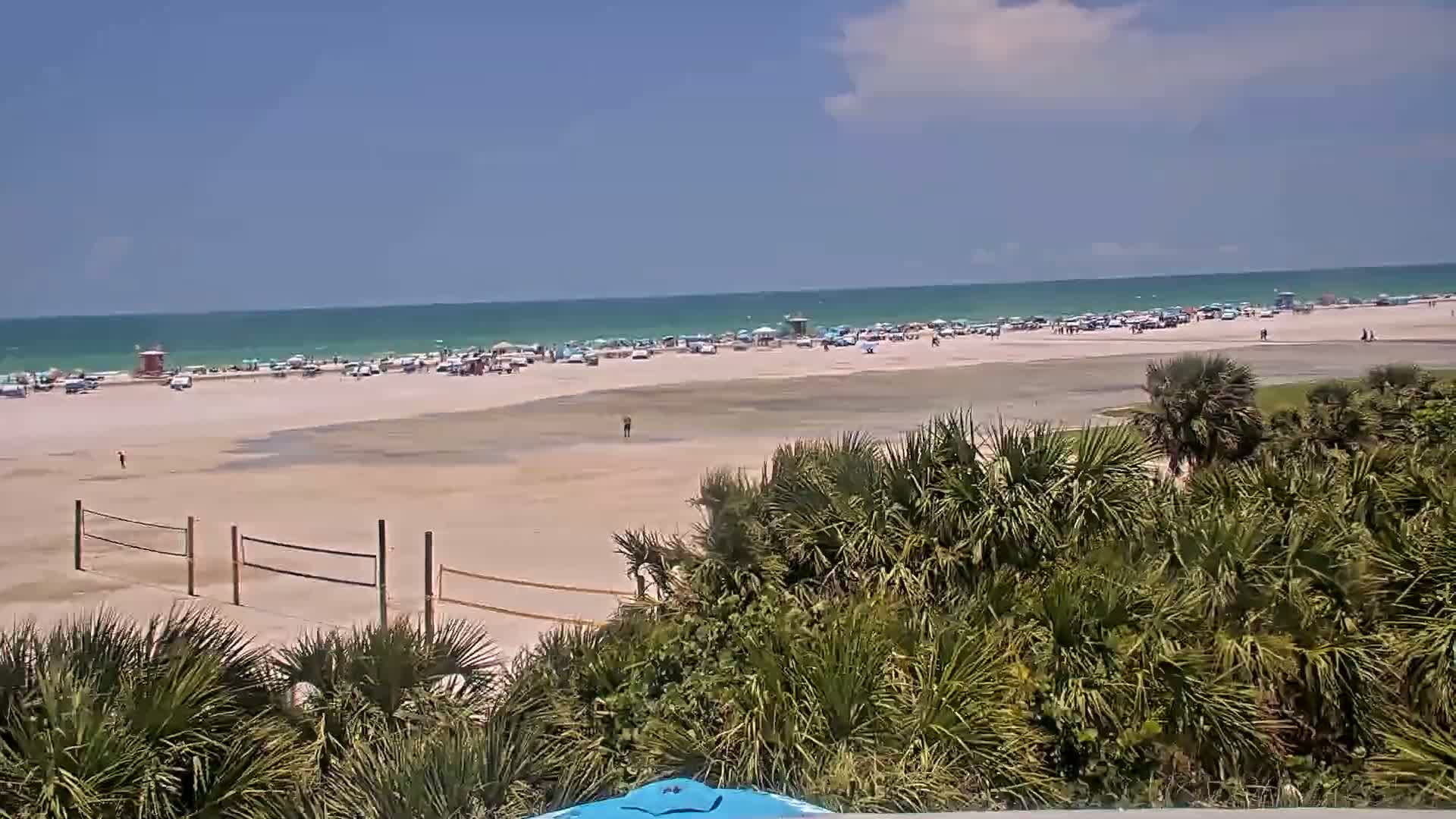 A sandy beach with a moderately populated shoreline, volleyball nets, and tropical vegetation is seen under sunny skies and calm ocean waters.