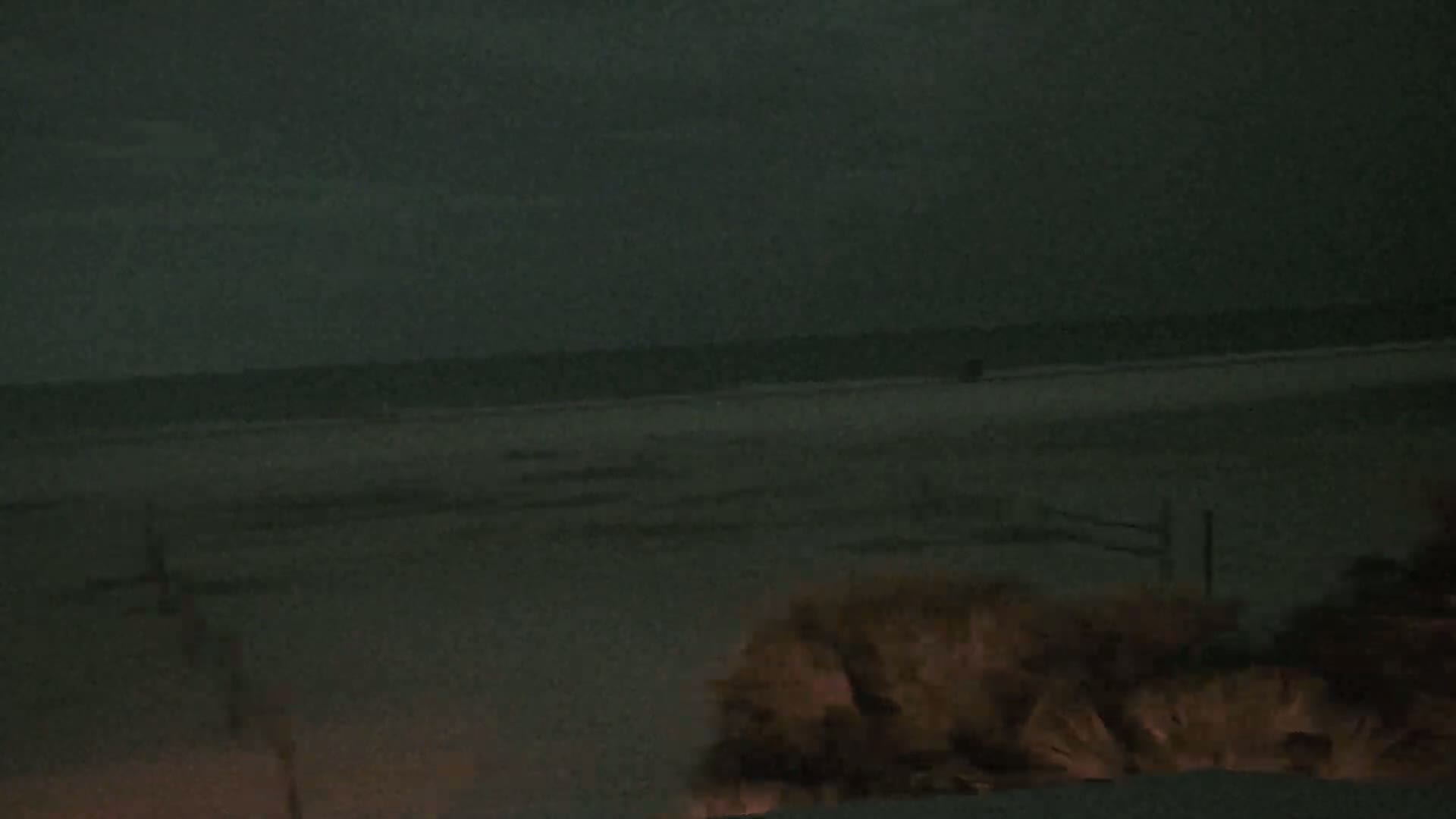 A dark, nighttime beach scene shows a calm, dark ocean under a cloudy, dark sky, with vegetation in the foreground.