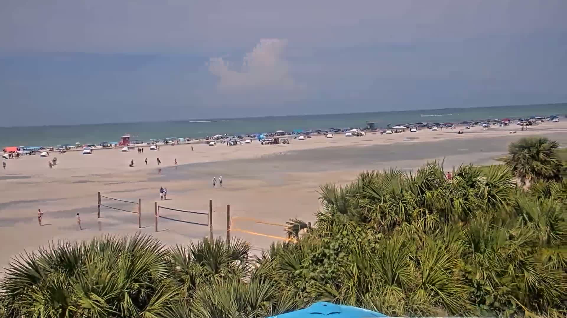A mostly sunny day at a beach shows a wide expanse of sand with scattered people, beach umbrellas, and a volleyball net, viewed from behind a line of palm trees.