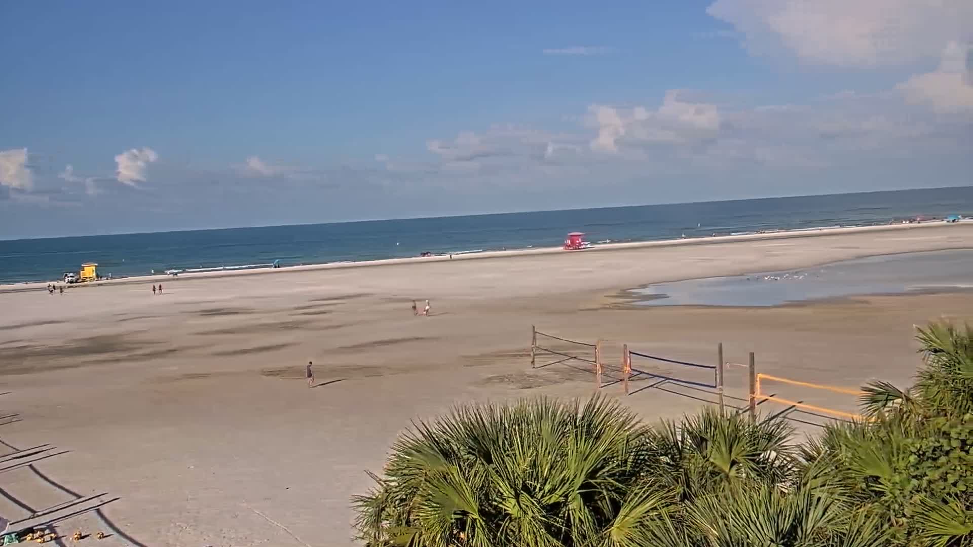 A mostly empty, sandy beach with a calm ocean under a partly cloudy sky features volleyball nets and lifeguard stands.