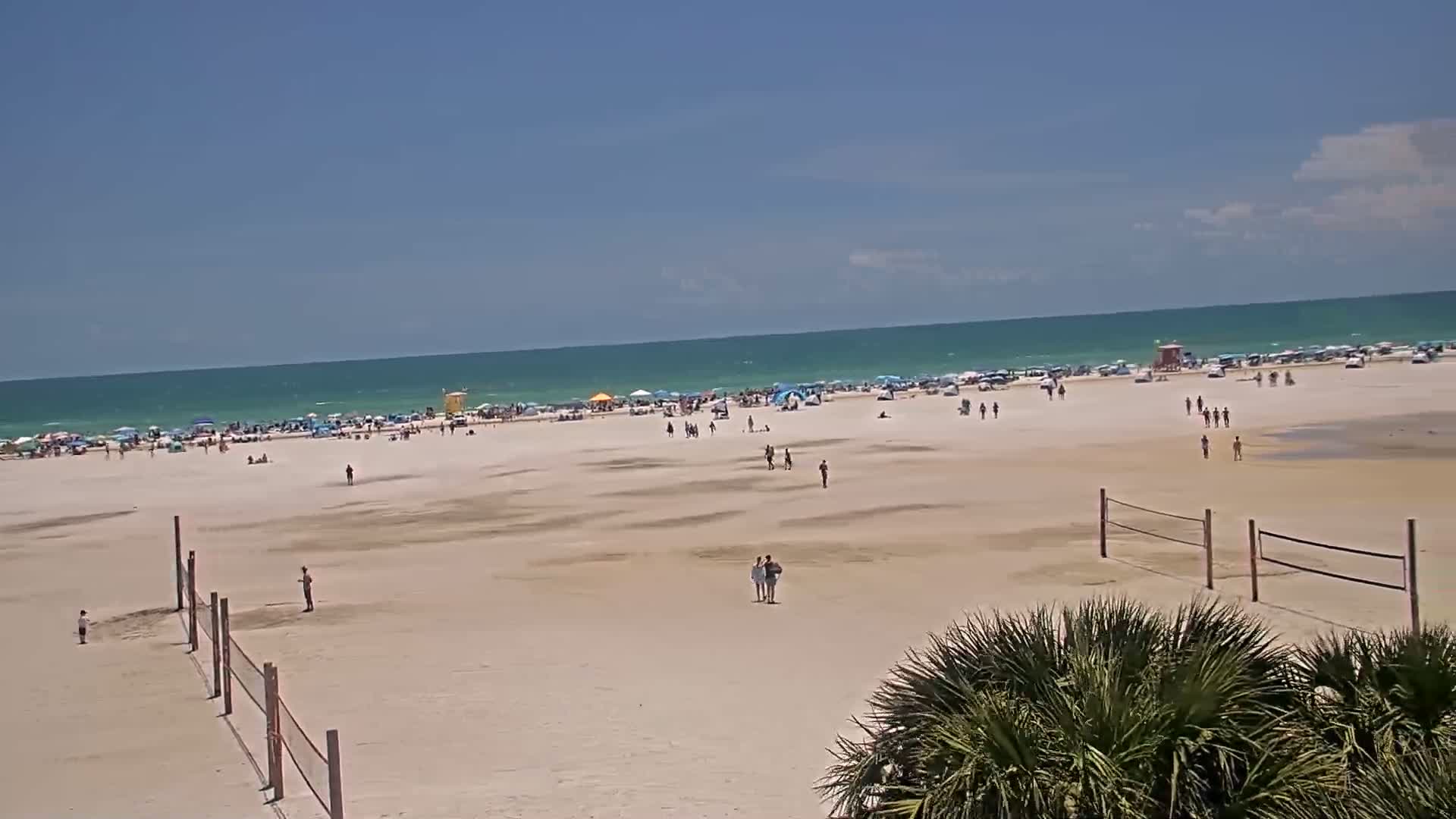 A mostly sunny day at a beach shows many people scattered across the sand and near the water, with volleyball nets in the foreground.