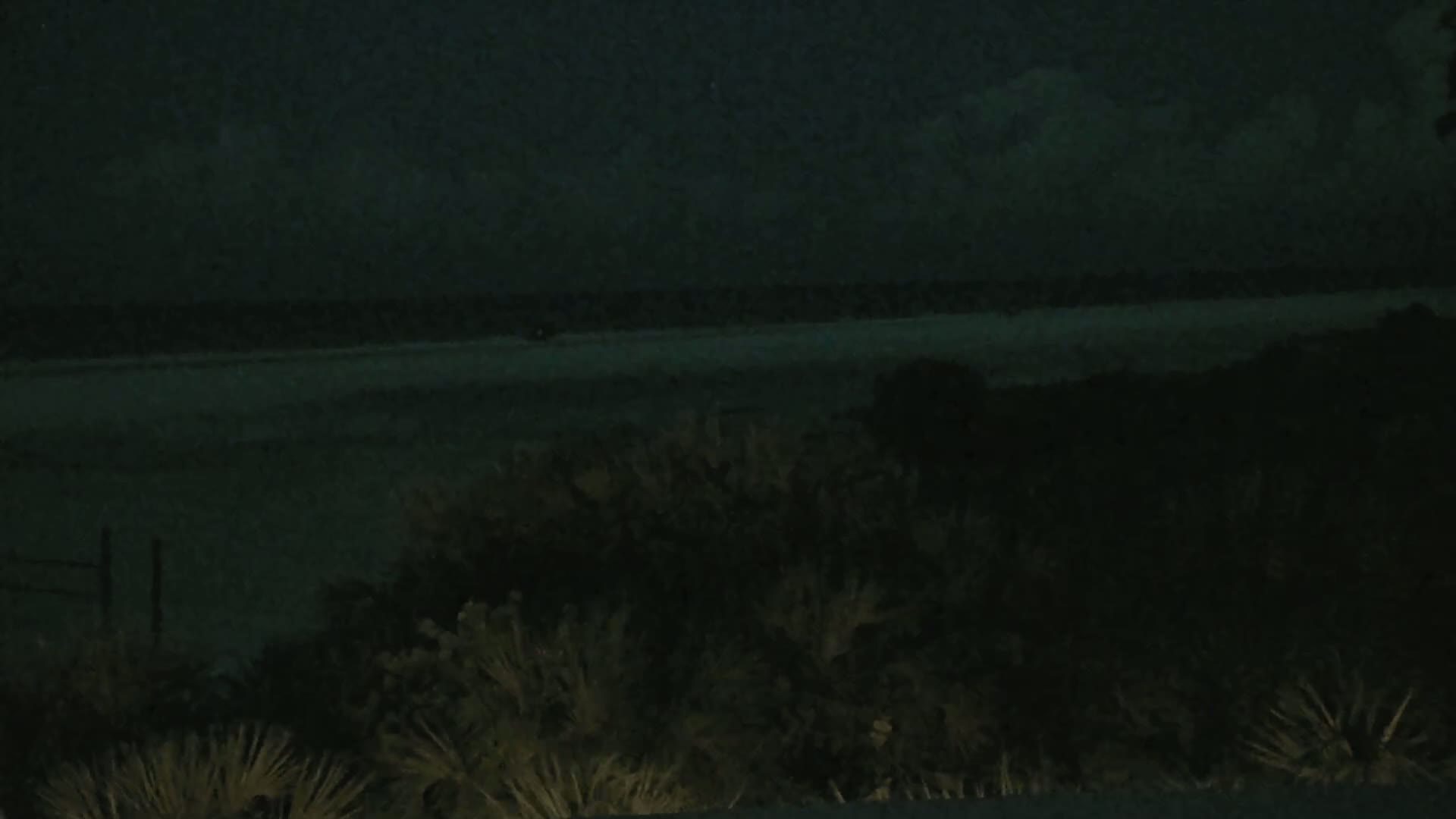 A dark, nighttime view shows a body of water in the distance, obscured by low-hanging clouds, with dark vegetation in the foreground.