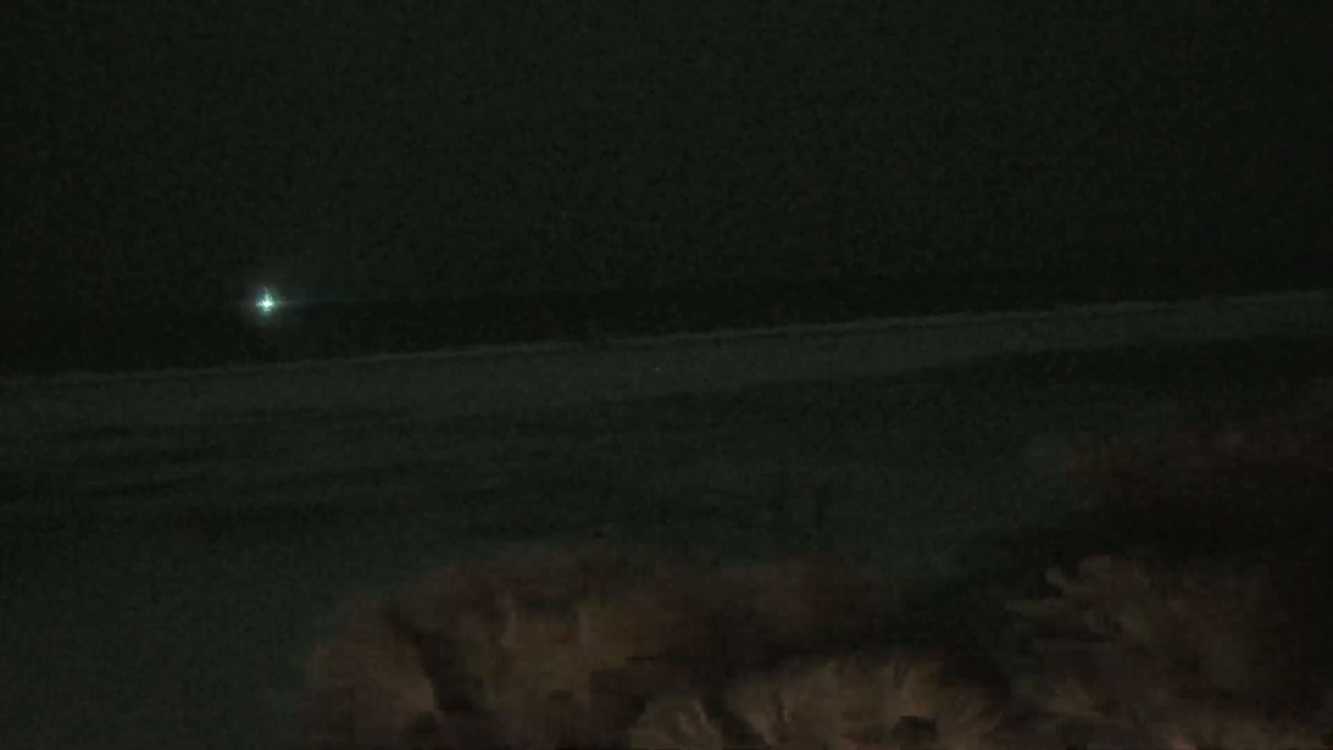 A dark, nighttime scene shows a faint light on the horizon above a dark beach and vegetation in the foreground.