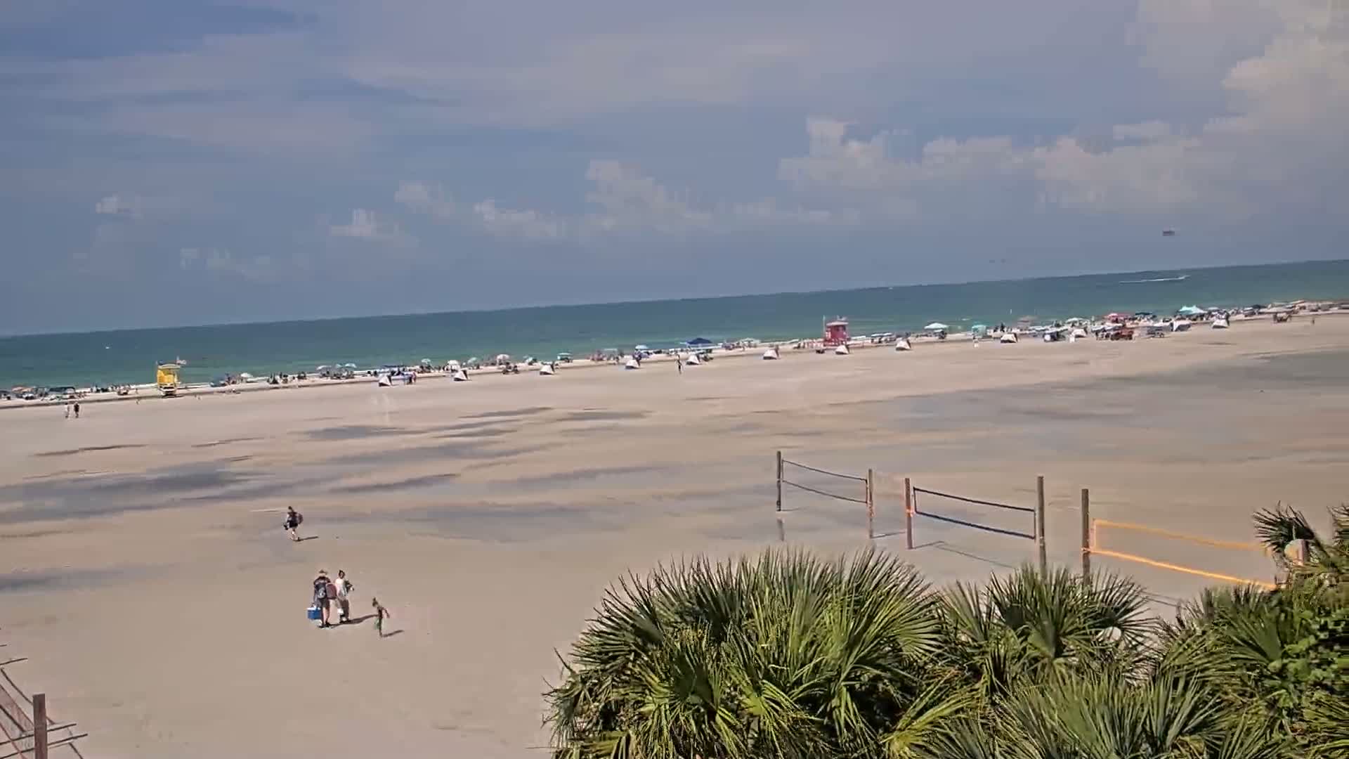 A mostly sunny beach scene shows a wide expanse of wet sand with scattered people, volleyball nets, and lifeguard stands, all under a partly cloudy sky.