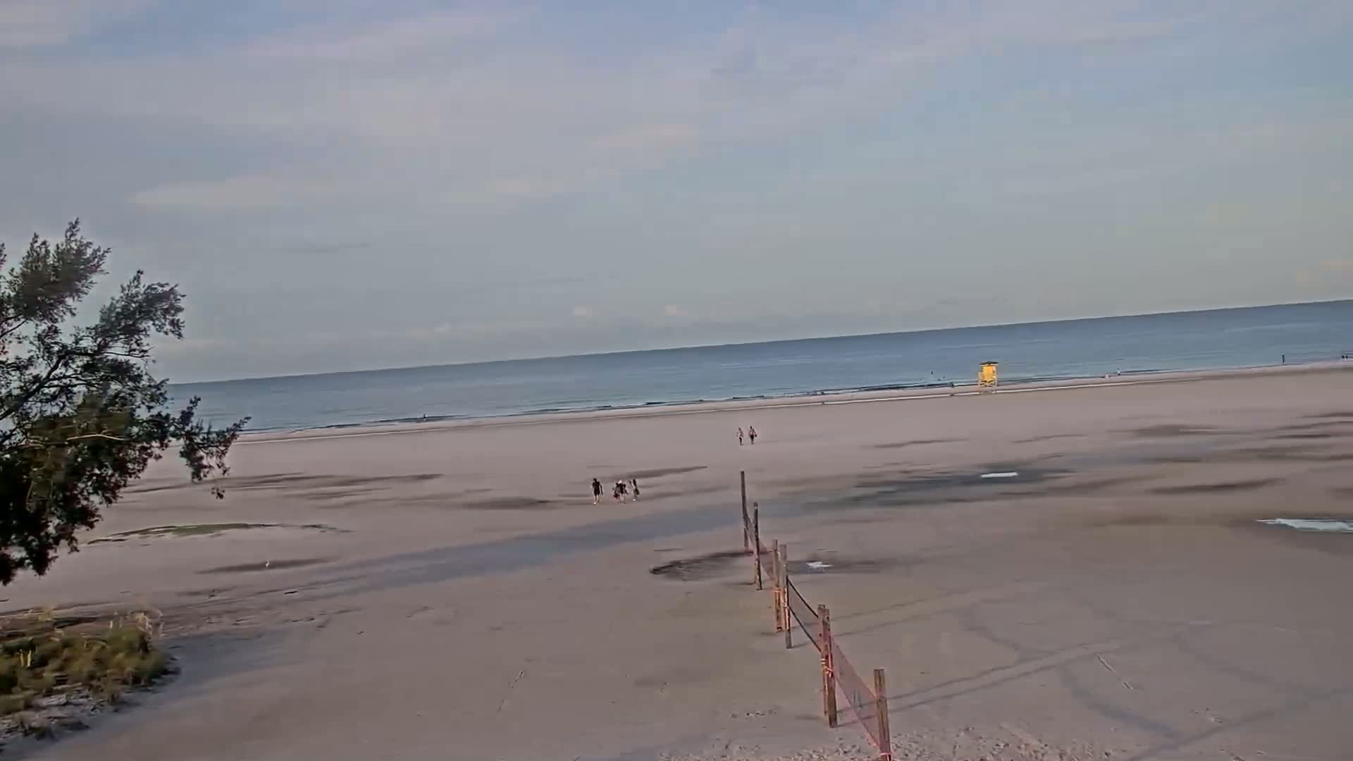 A mostly empty beach with a few people and a lifeguard stand is visible under a mostly cloudy sky, with a calm ocean in the background.