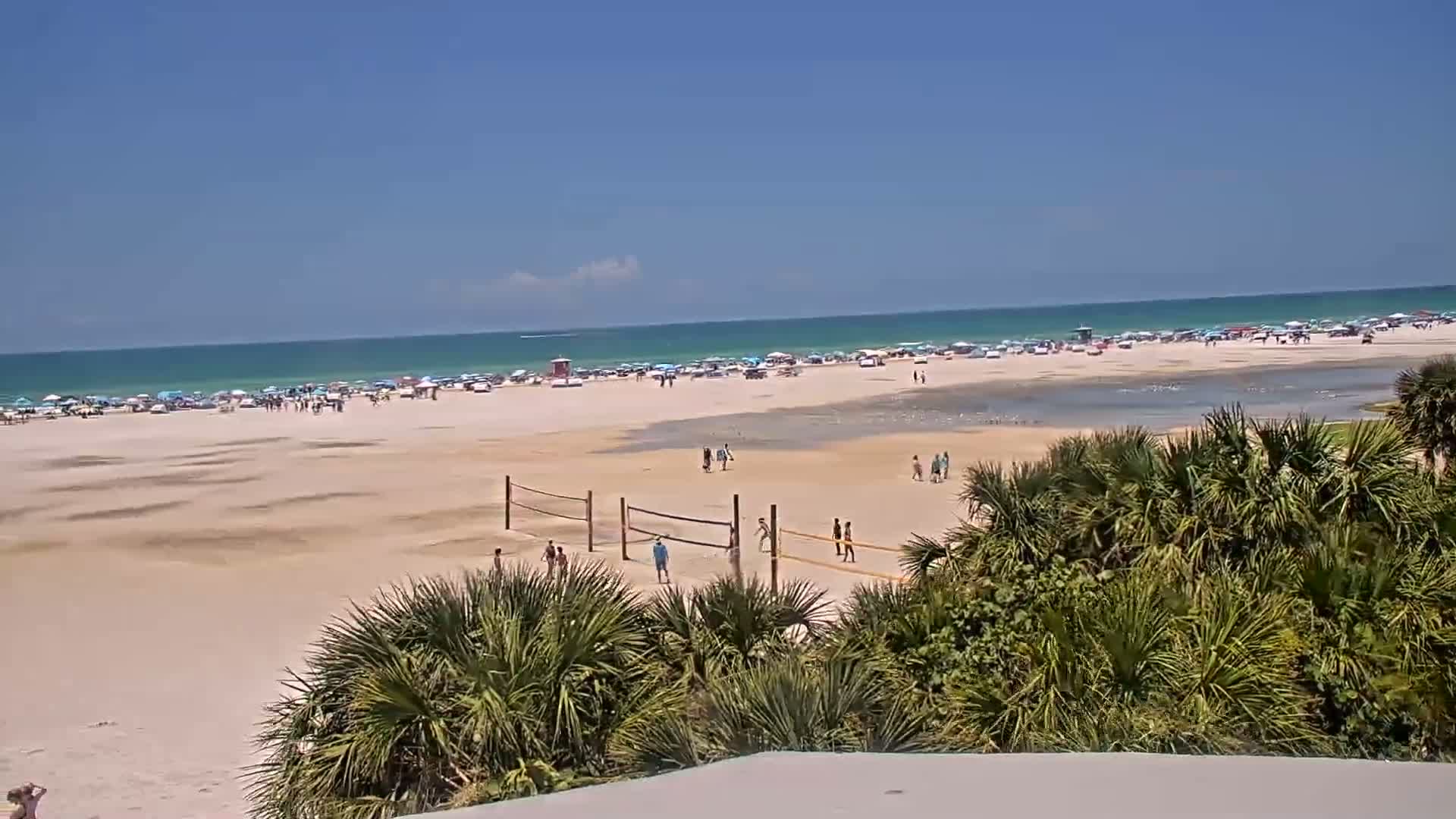 A sunny beach scene shows a wide expanse of sand with people, beach umbrellas, and volleyball nets, all under a clear blue sky and calm ocean.