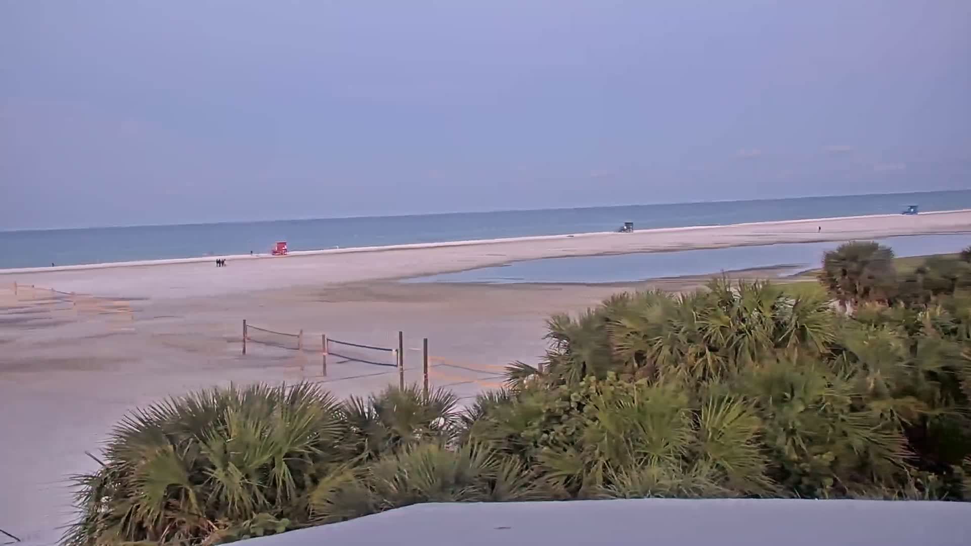 A mostly empty beach with calm water, volleyball nets, and palm trees in the foreground under a clear sky.