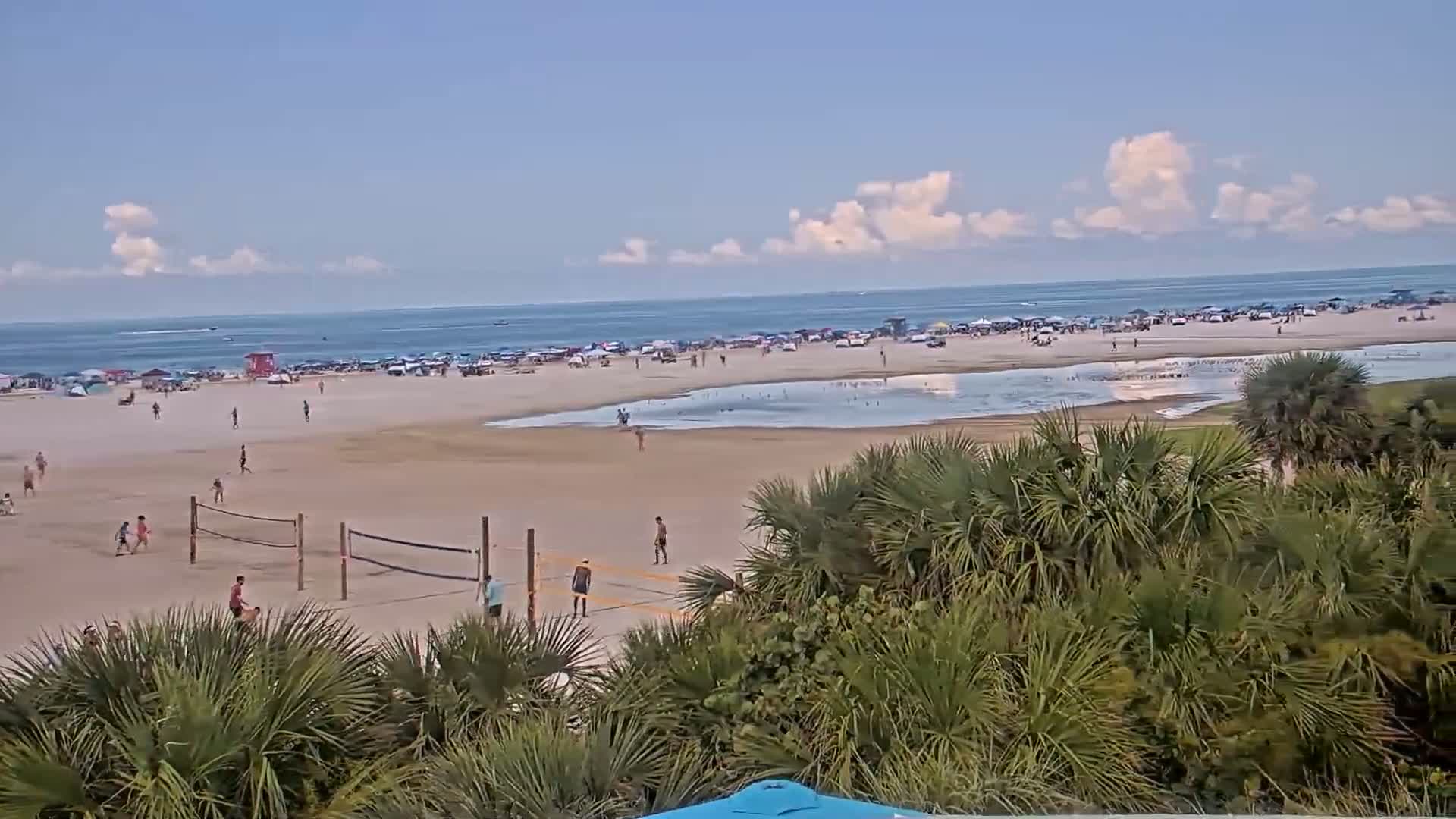 A sandy beach with numerous people, some playing volleyball, is partially obscured by palm trees under a partly cloudy sky.