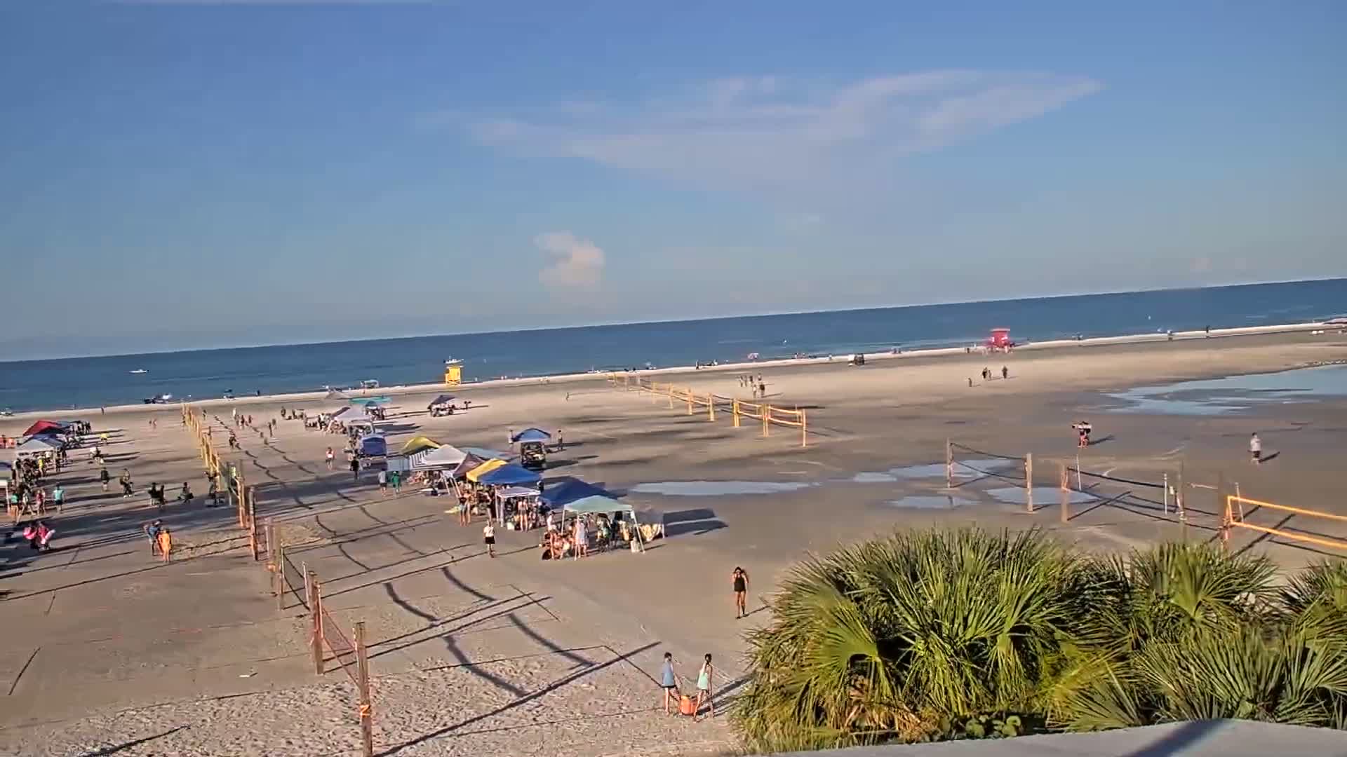 A sunny beach scene shows several volleyball nets set up on the sand, with people playing and relaxing under various tents and umbrellas, near a calm ocean.