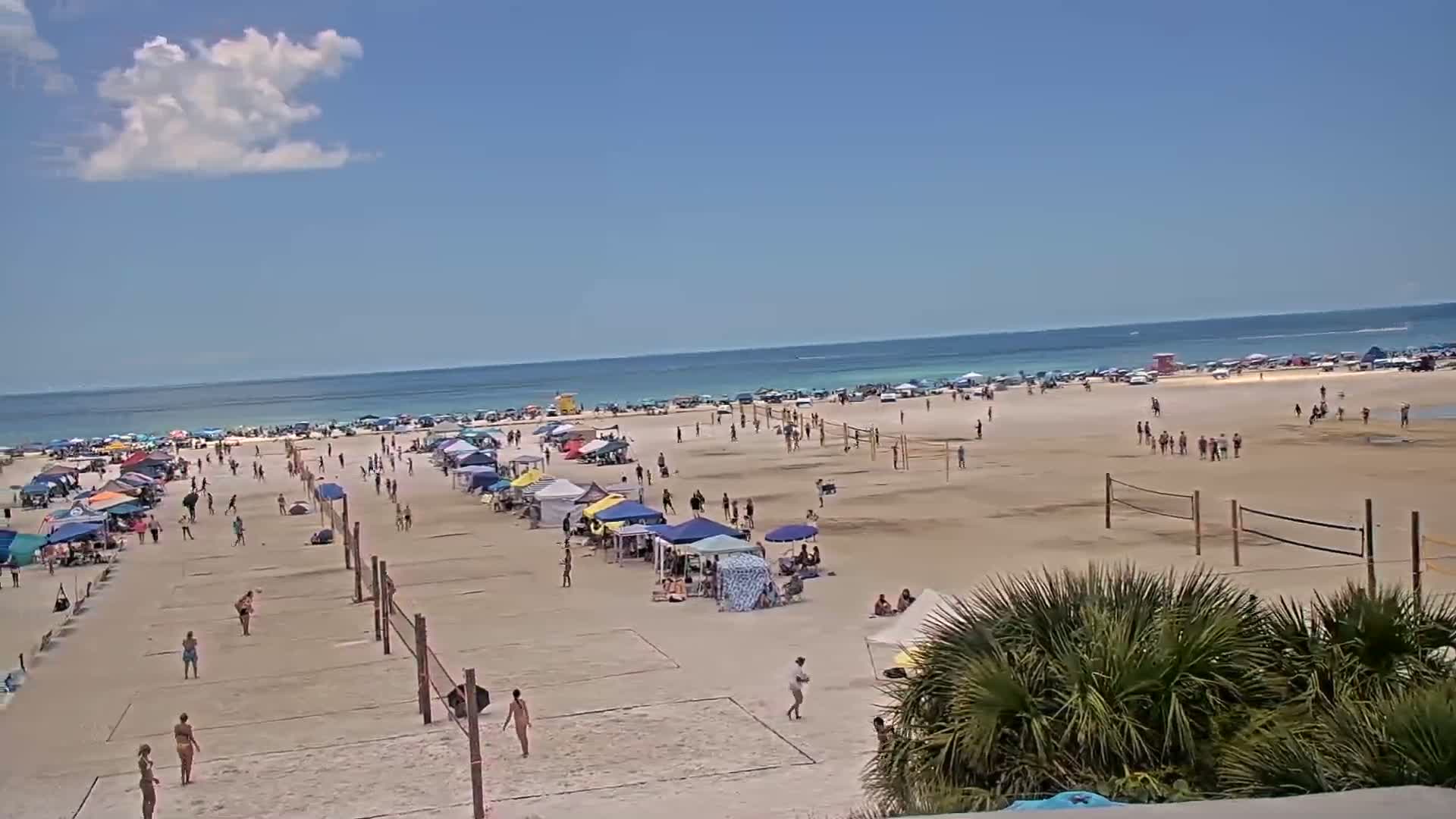 A sandy beach with numerous people, volleyball nets, and beach umbrellas under a partly cloudy sky and calm ocean.