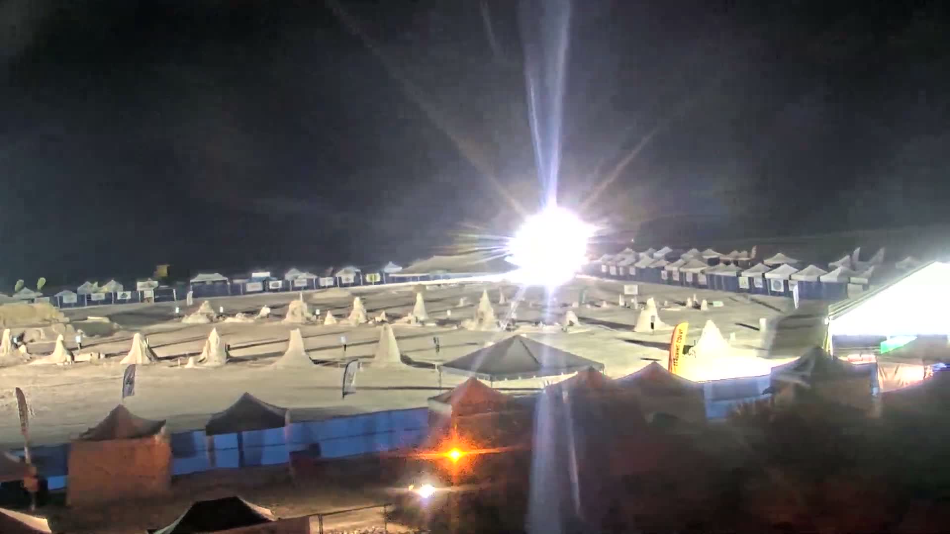 The image displays a sprawling outdoor encampment at night, featuring numerous tents and conical structures on sandy ground, brightly illuminated by a powerful, flaring light source under a clear dark sky.