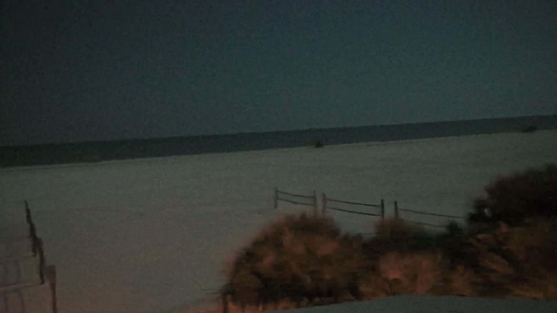 A calm, dimly lit beach scene at twilight or night features a vast expanse of pale sand leading to a dark ocean horizon, with a faint wooden fence and shadowy foreground foliage under a deep blue-grey sky.