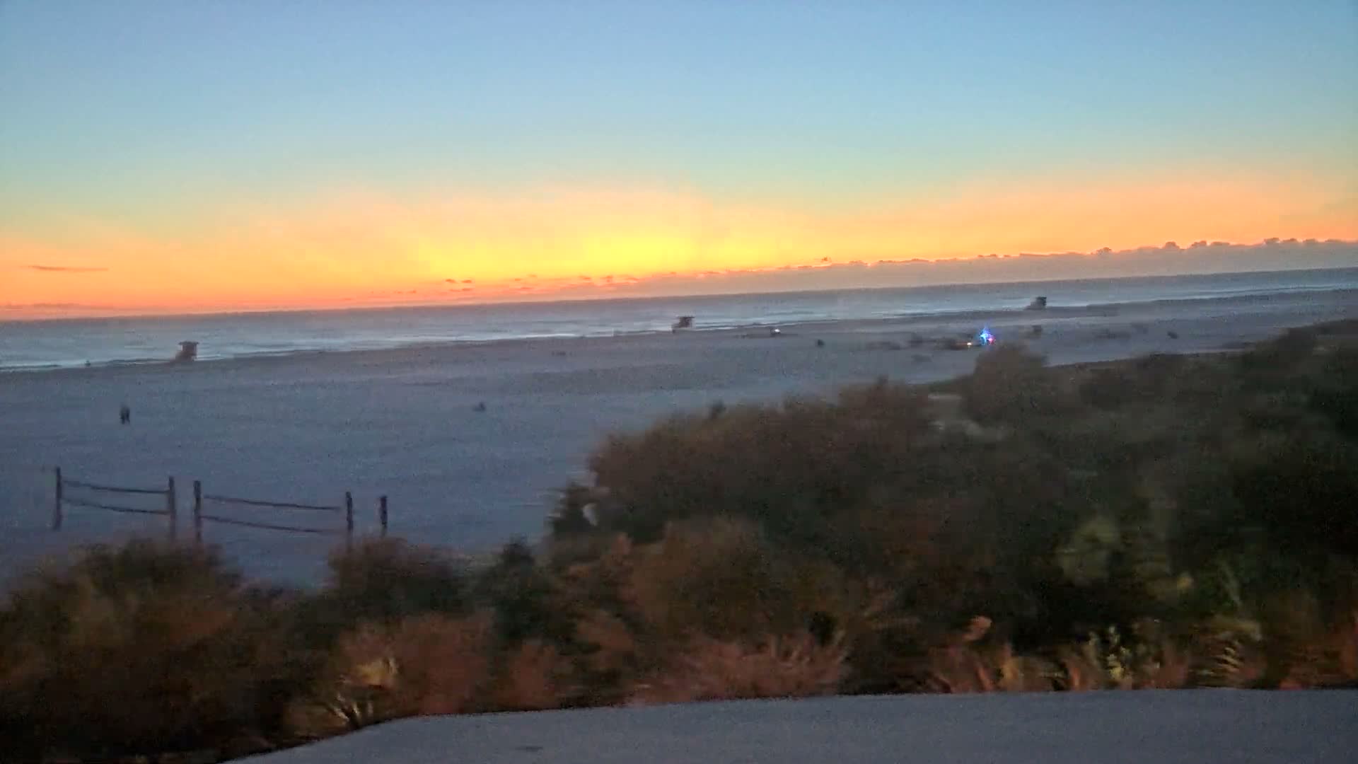 A calm, dimly lit beach scene at twilight or night features a vast expanse of pale sand leading to a dark ocean horizon, with a faint wooden fence and shadowy foreground foliage under a deep blue-grey sky.