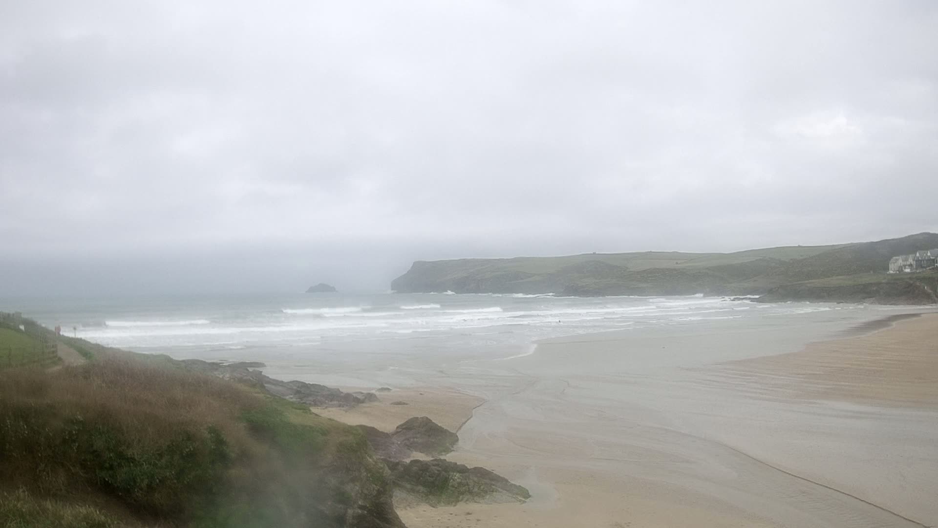 A hazy, overcast day reveals a sandy beach meeting a choppy ocean, backed by green hills and a distant island.