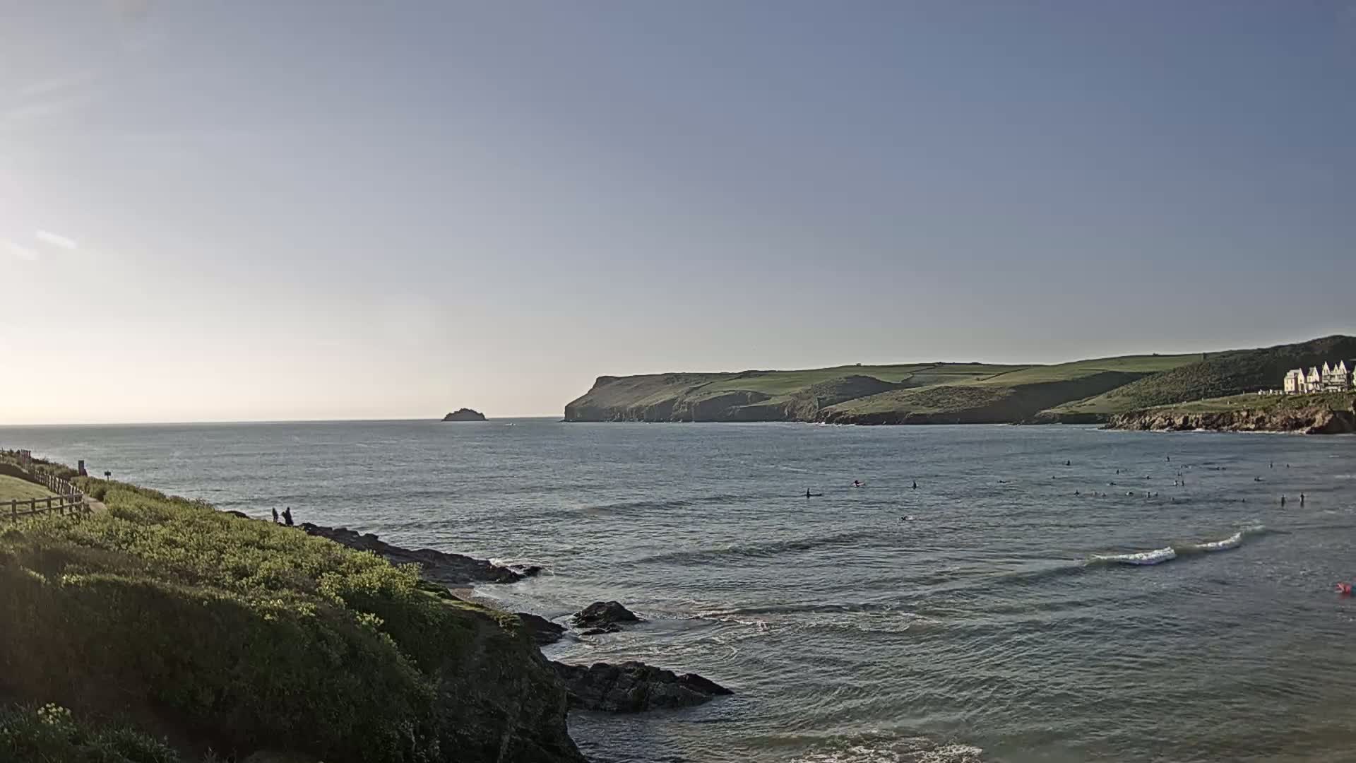 A calm ocean bay with small waves, surfers, and a distant island under a clear sky is visible from a green, rocky cliffside.