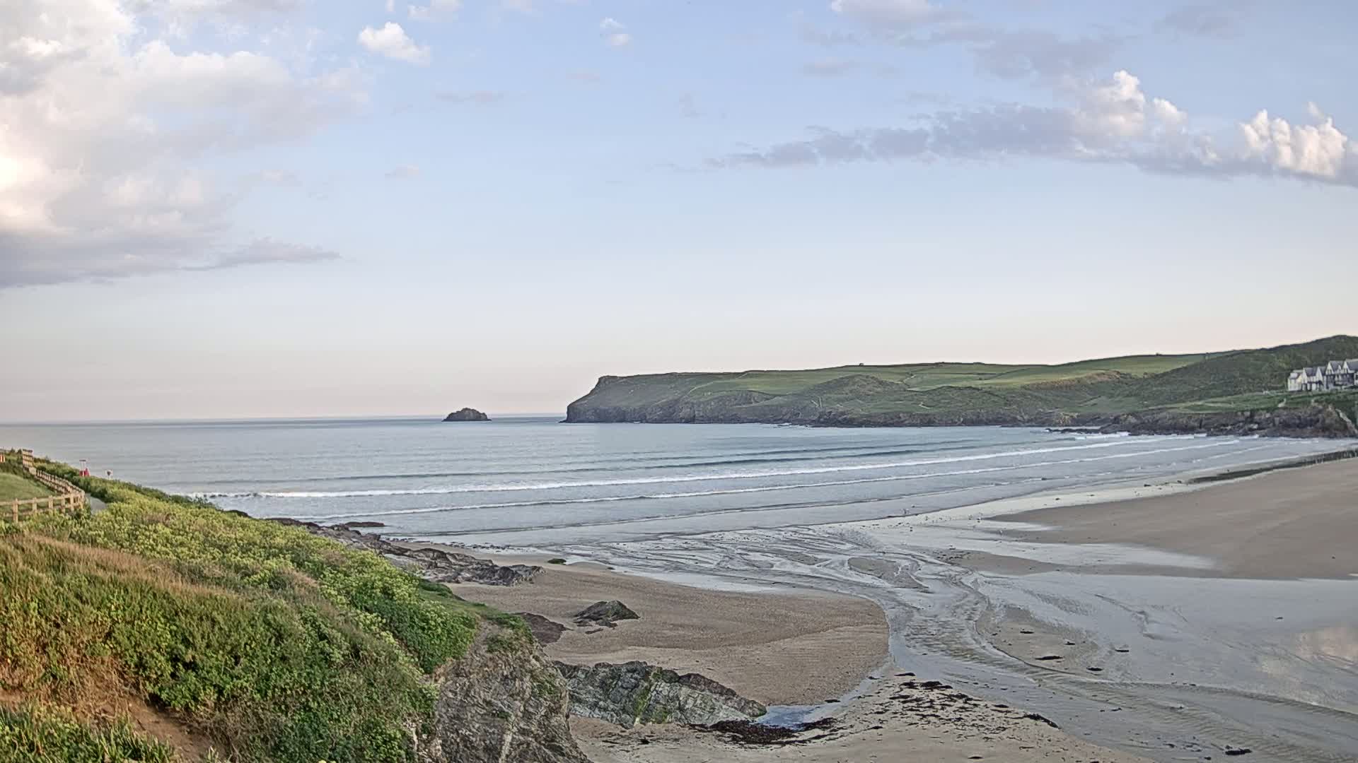 A sandy beach with gentle waves stretches out to meet a calm ocean under a partly cloudy sky, with a small island visible in the distance and green hills lining the coast.