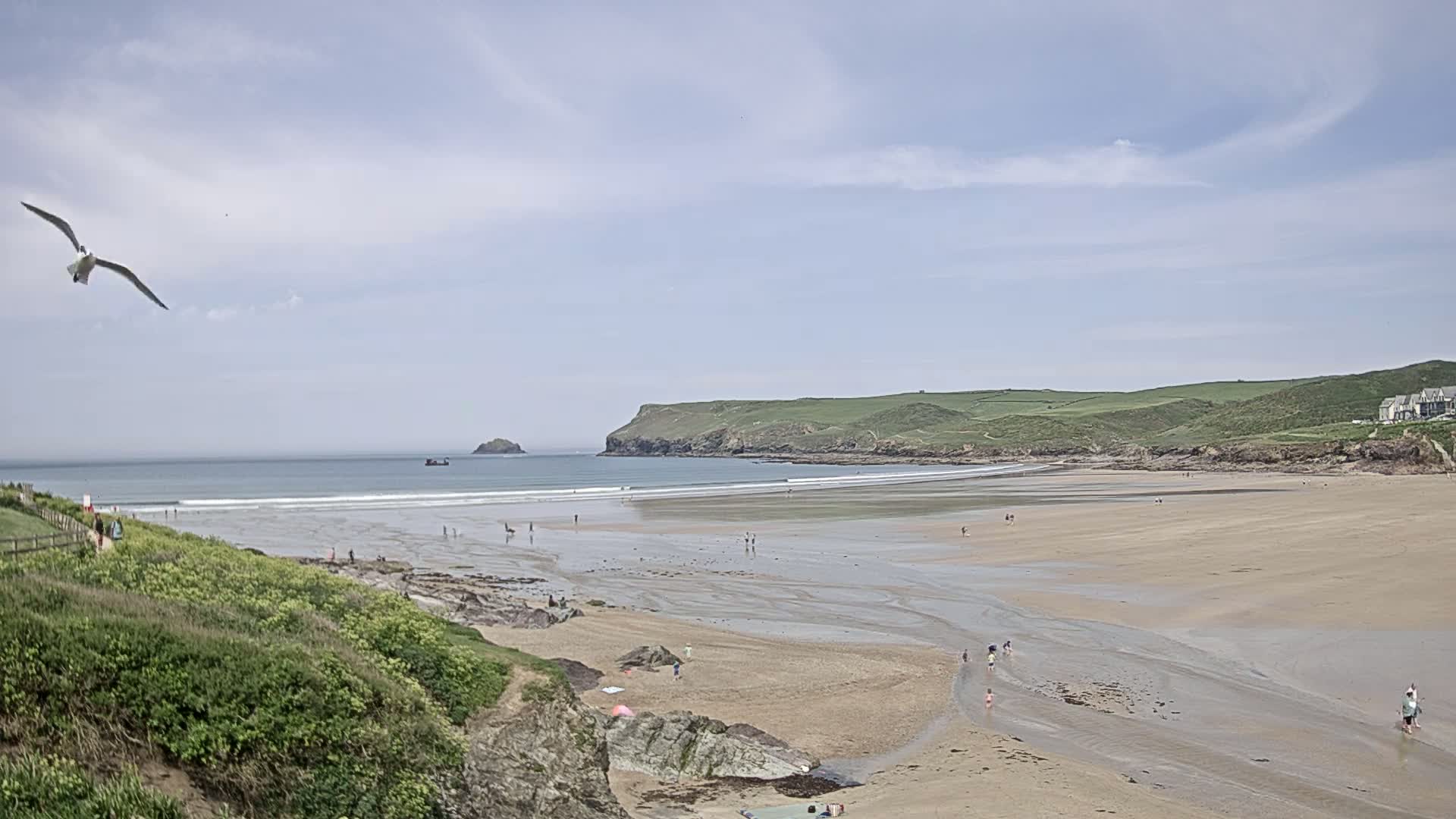 A seagull flies over a sandy beach with a few people scattered across it, under a partly cloudy sky, with green hills and a distant island in the background.