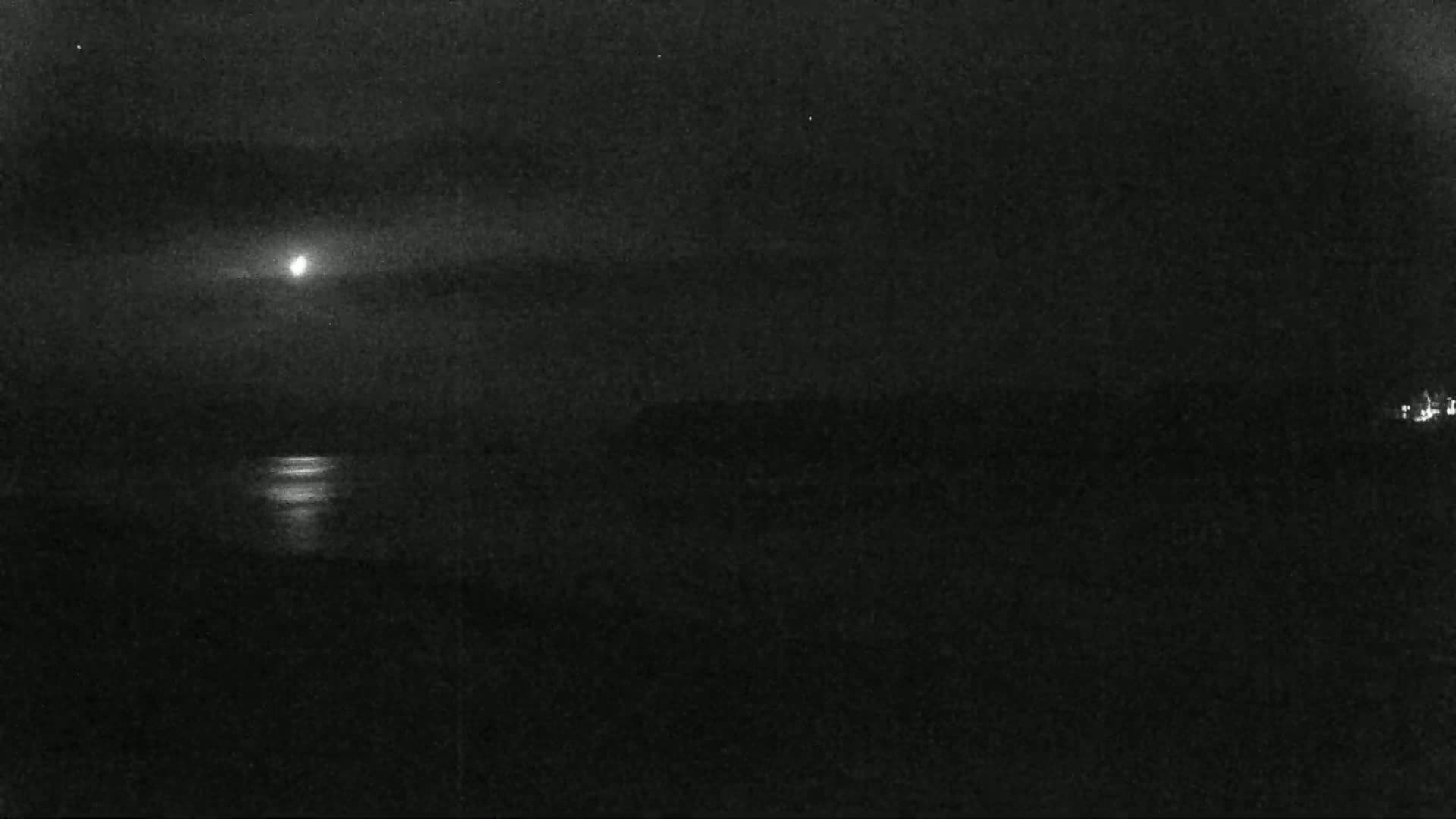 A bright moon shines over dark water reflecting its light, with a dark landmass in the distance under a mostly clear night sky.