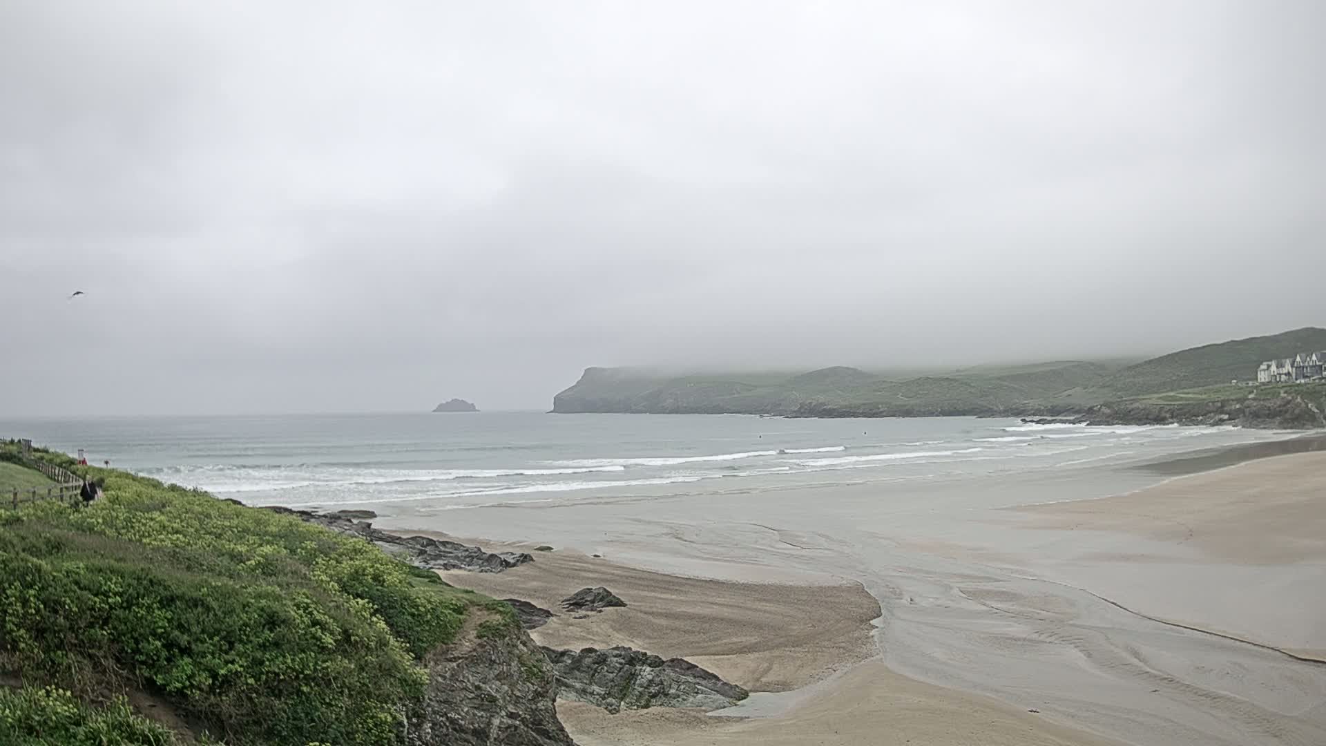 A wide sandy beach stretches out to a calm, grey ocean under an overcast sky, with green hills and a distant island in the background.