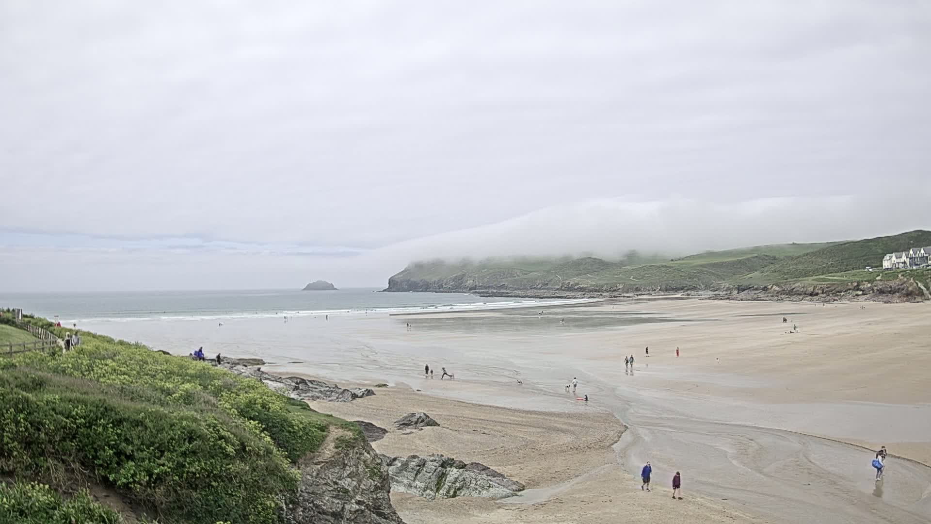 A wide sandy beach stretches to a calm ocean under a cloudy sky, with a few people scattered across the sand and several more in the water.