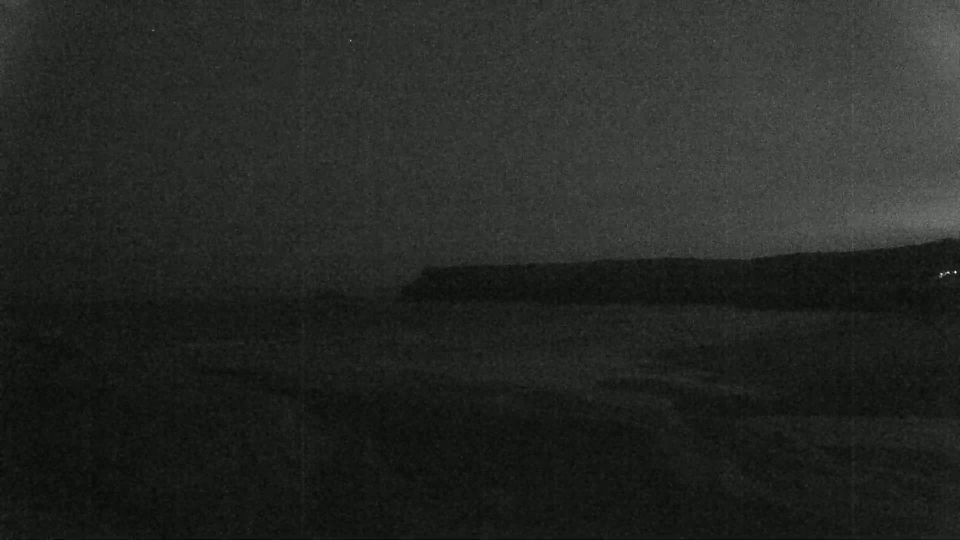 A dark, nighttime coastal scene shows a dark shoreline under a mostly clear sky.