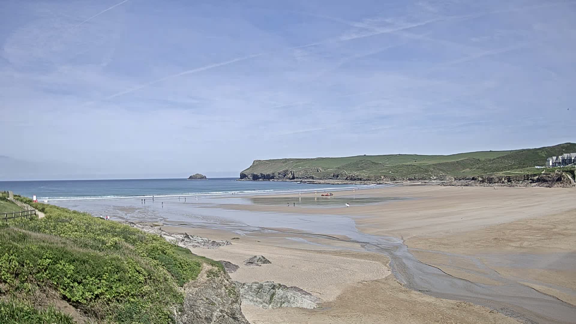 A sandy beach with small waves and a few people stretches out under a clear, bright sky, backed by green hills and a distant headland.