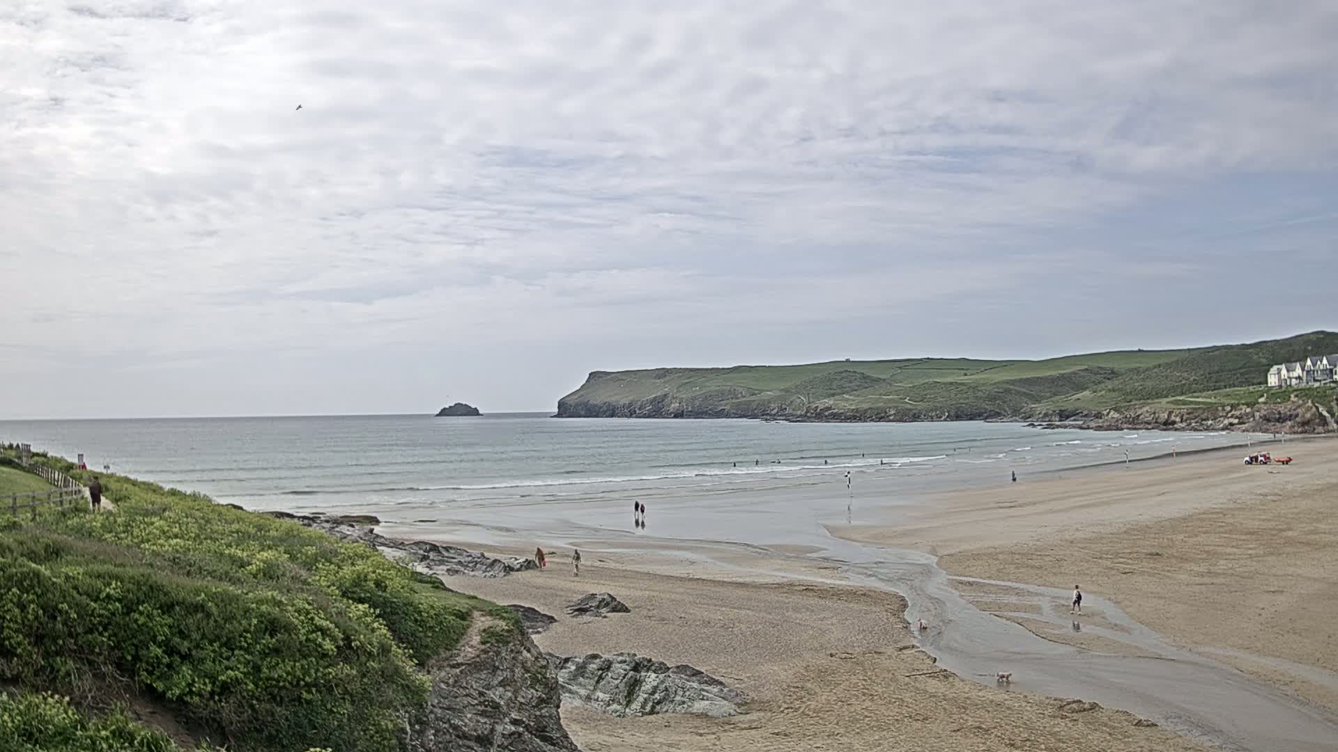 A sandy beach with a few people and a small stream flows into the ocean under a mostly cloudy sky.