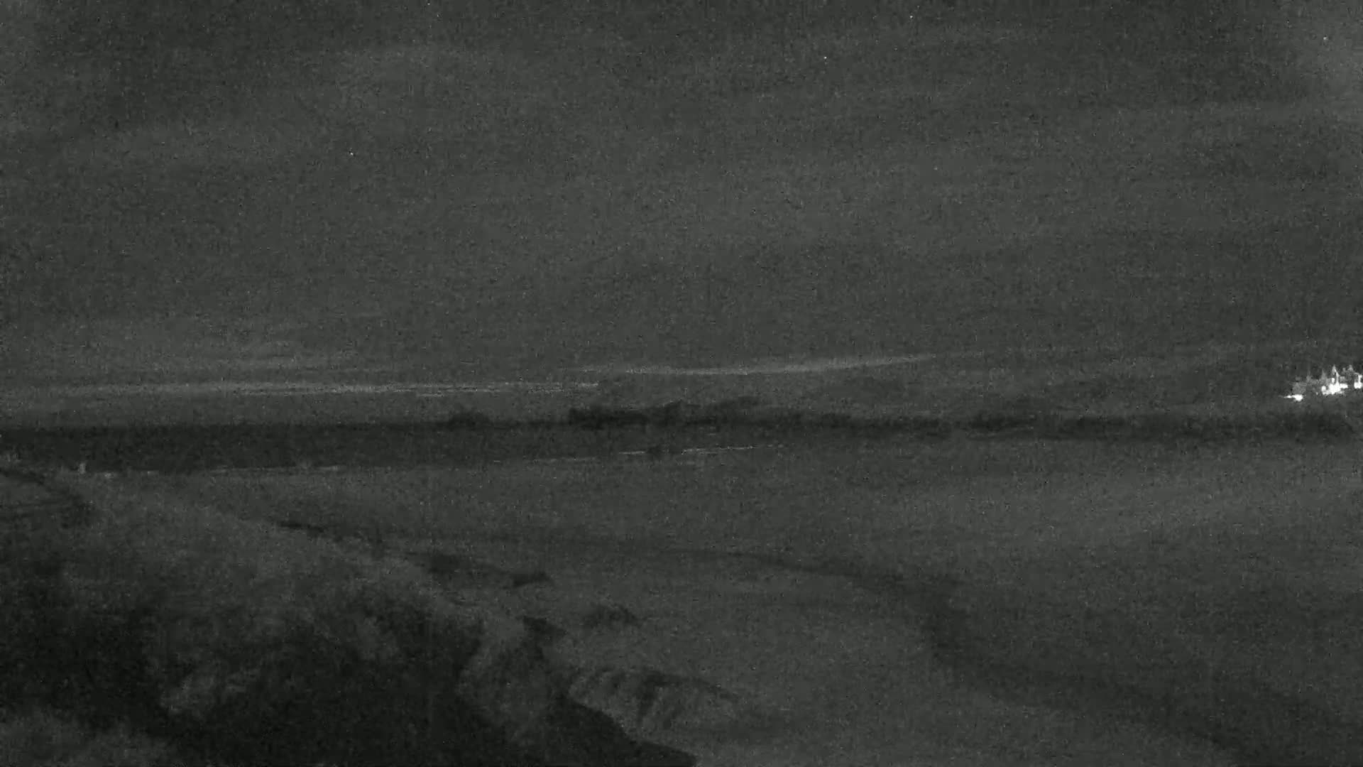A dark, nighttime view shows a flat expanse of land meeting a calm body of water under a cloudy sky.