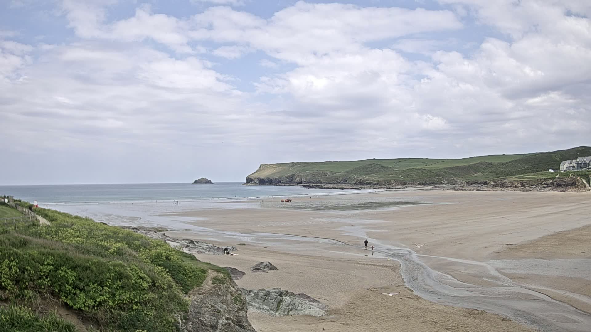A sandy beach stretches out to a calm ocean under a partly cloudy sky, with a small stream cutting through the sand and green hills rising in the background.