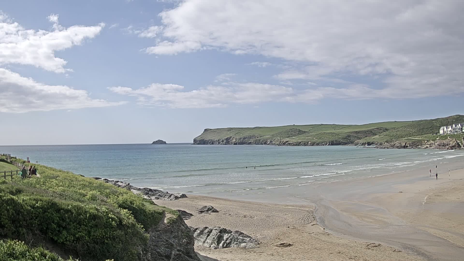 A sandy beach with gentle waves laps at the shore under a partly cloudy sky, with green hills and a small island in the distance.
