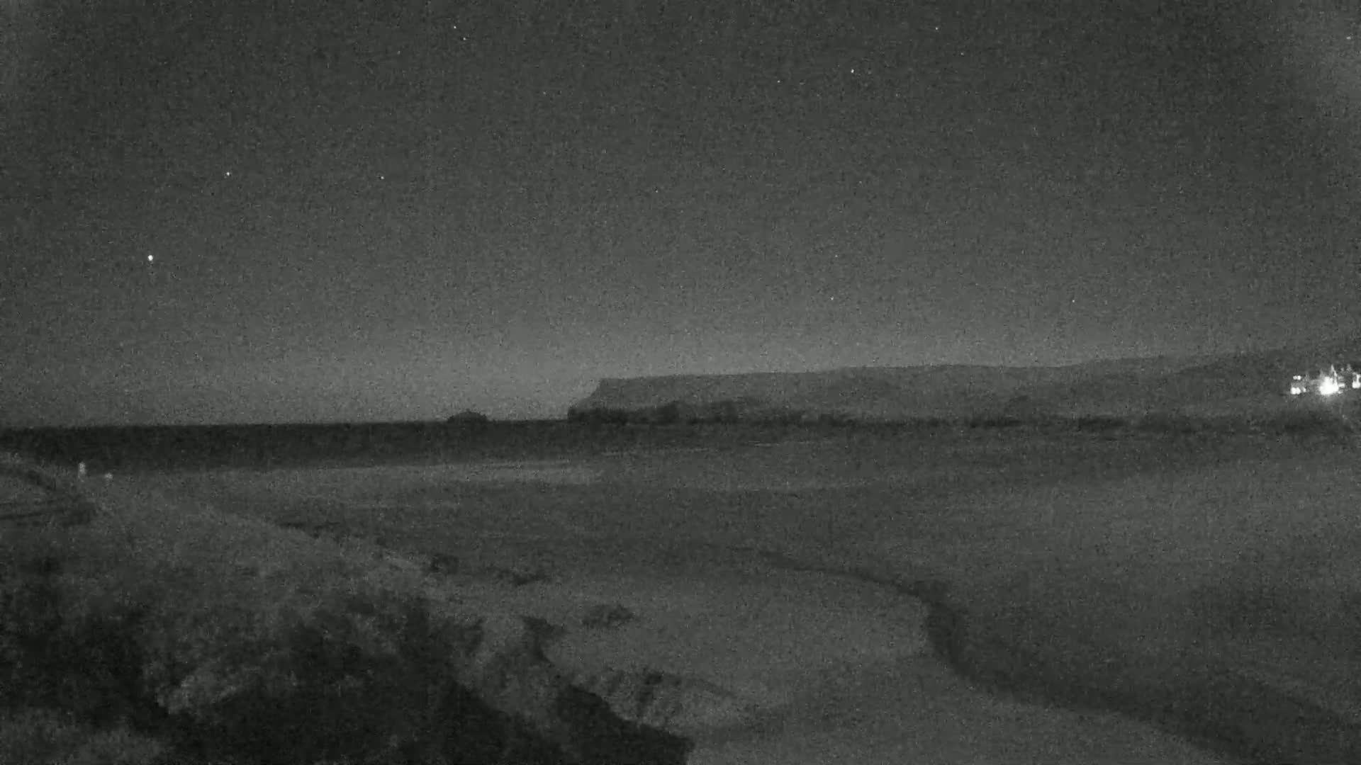 A dark, nighttime beach scene shows a calm ocean, a distant cliff line under a mostly clear, star-studded sky.