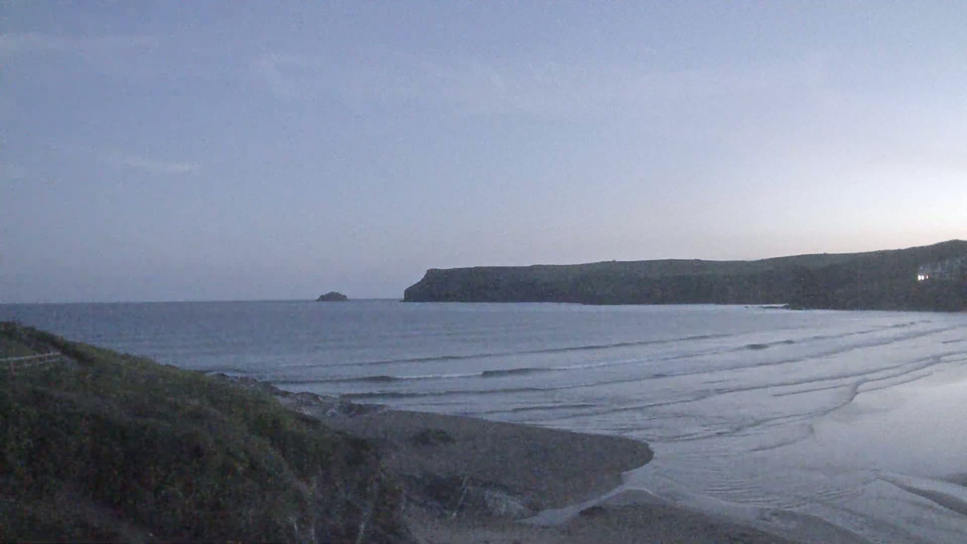 A calm, twilight ocean scene shows a sandy beach, small waves, a rocky headland, and a distant island under a clear, pale sky.