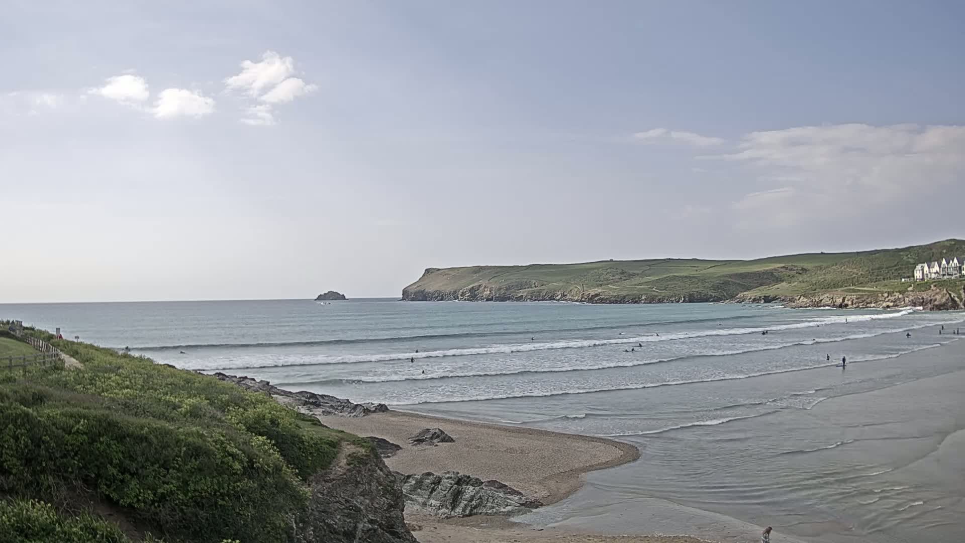 A sandy beach with gentle waves and several surfers is visible, under a partly cloudy sky.
