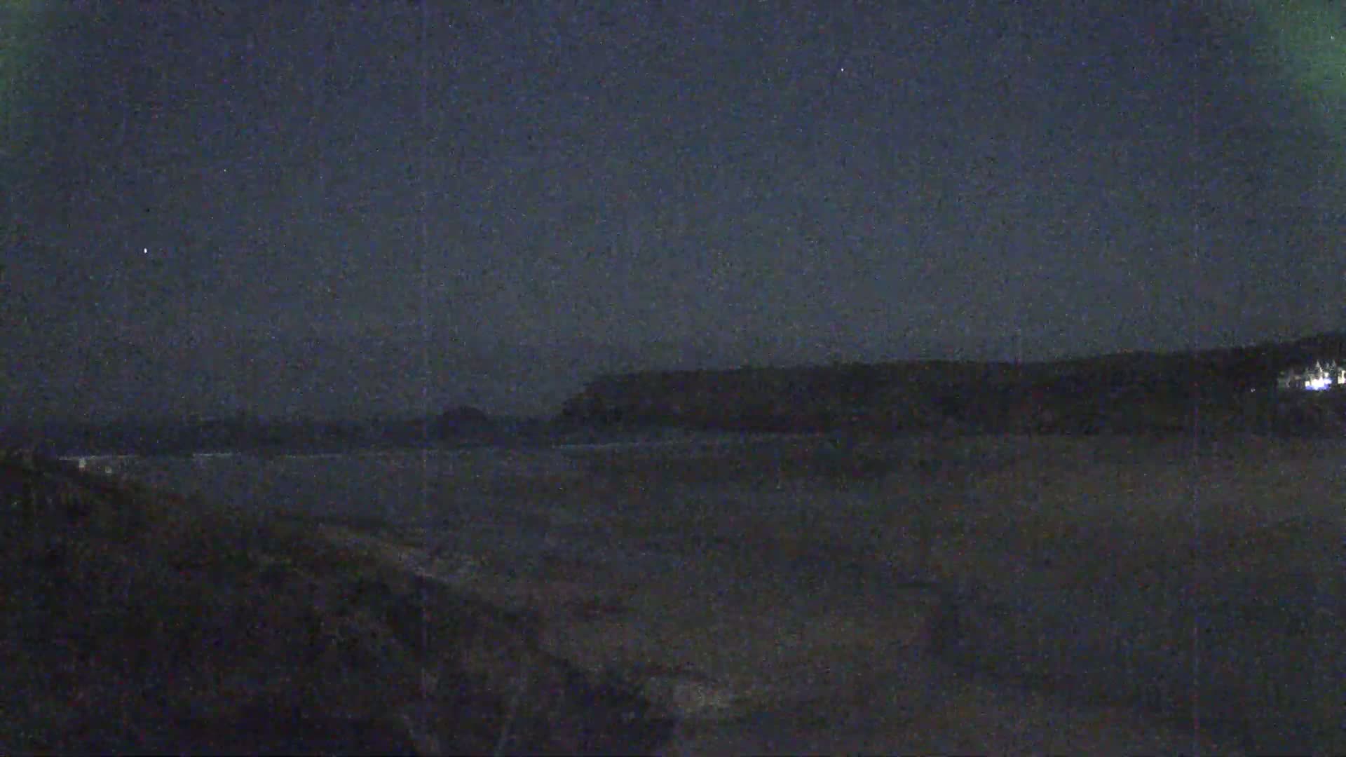 A dark, nighttime view of a beach with a distant, silhouetted shoreline under a mostly clear sky.