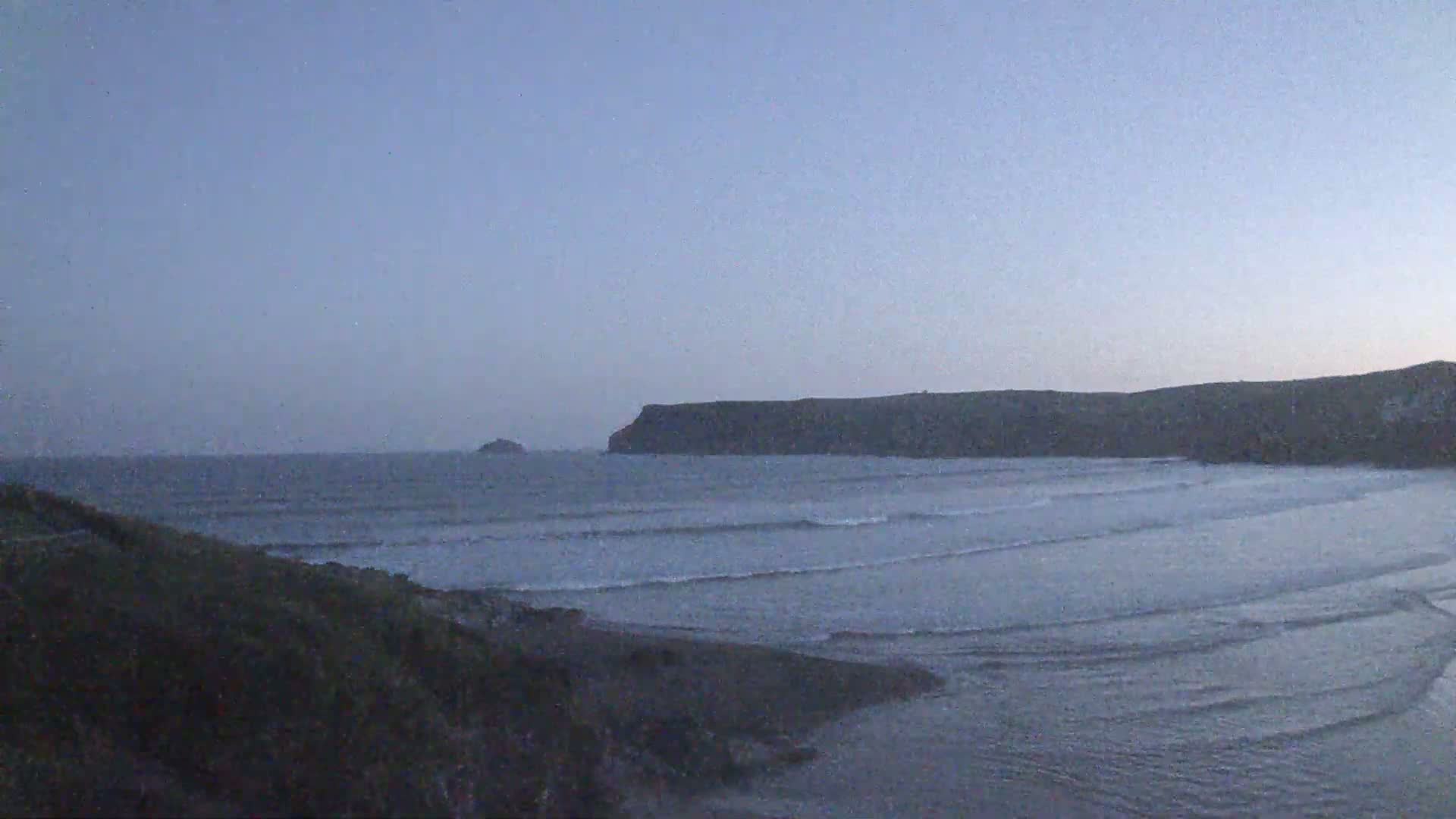 Under a clear, dusky sky, gentle waves lap a sandy beach, backed by a dark, rocky headland and a small island in the distance.