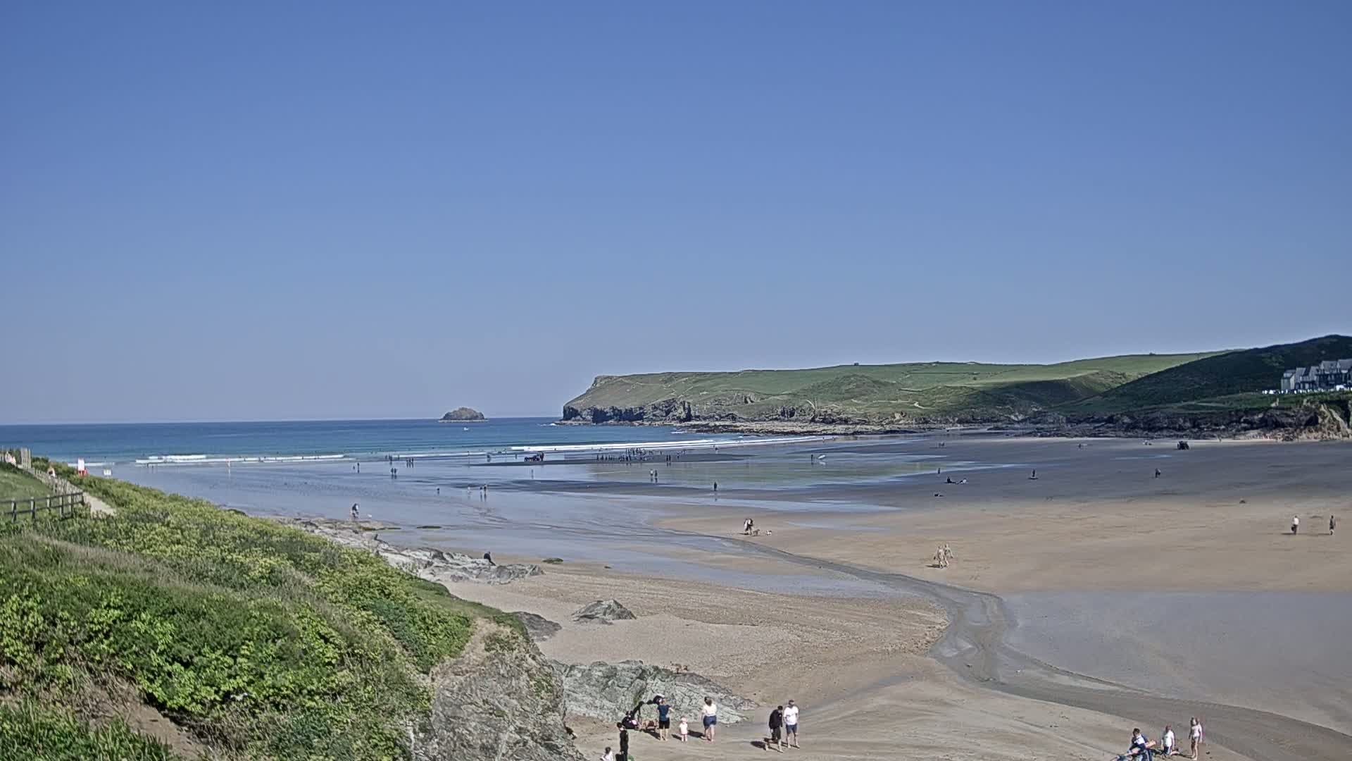 A sandy beach stretches out to a calm ocean under a clear blue sky, with people scattered across the sand and a few in the water.