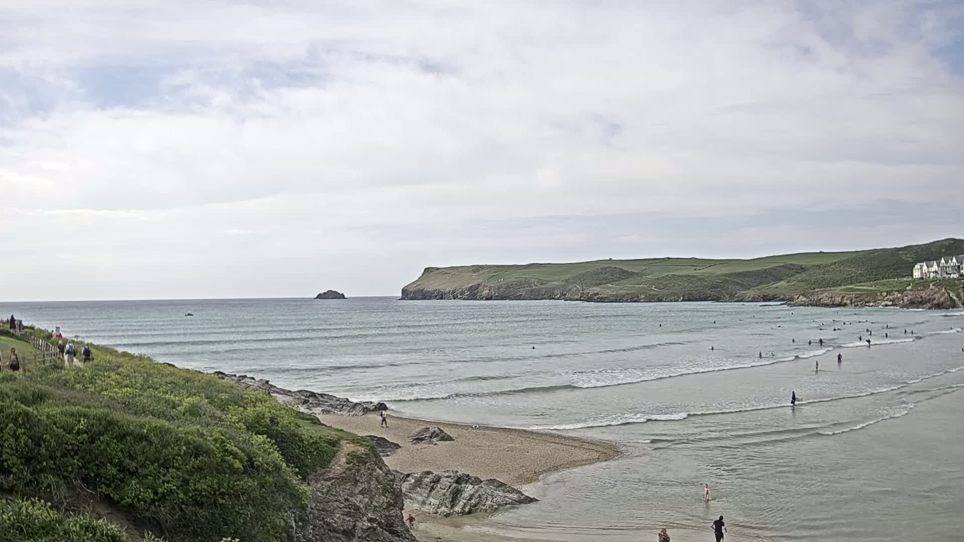 A sandy cove with gentle waves and numerous people swimming and surfing under an overcast sky.